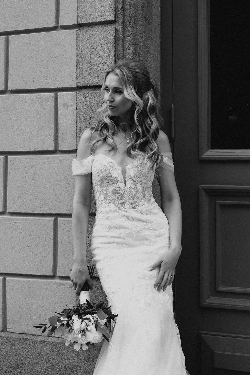 Black and white portrait of bride in off-shoulder lace gown holding bouquet beside stone building door.