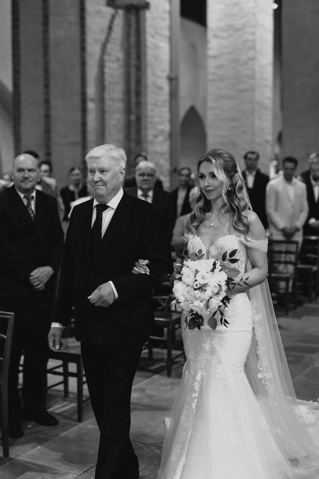 Bride in lace gown with veil walks down church aisle escorted by her father.