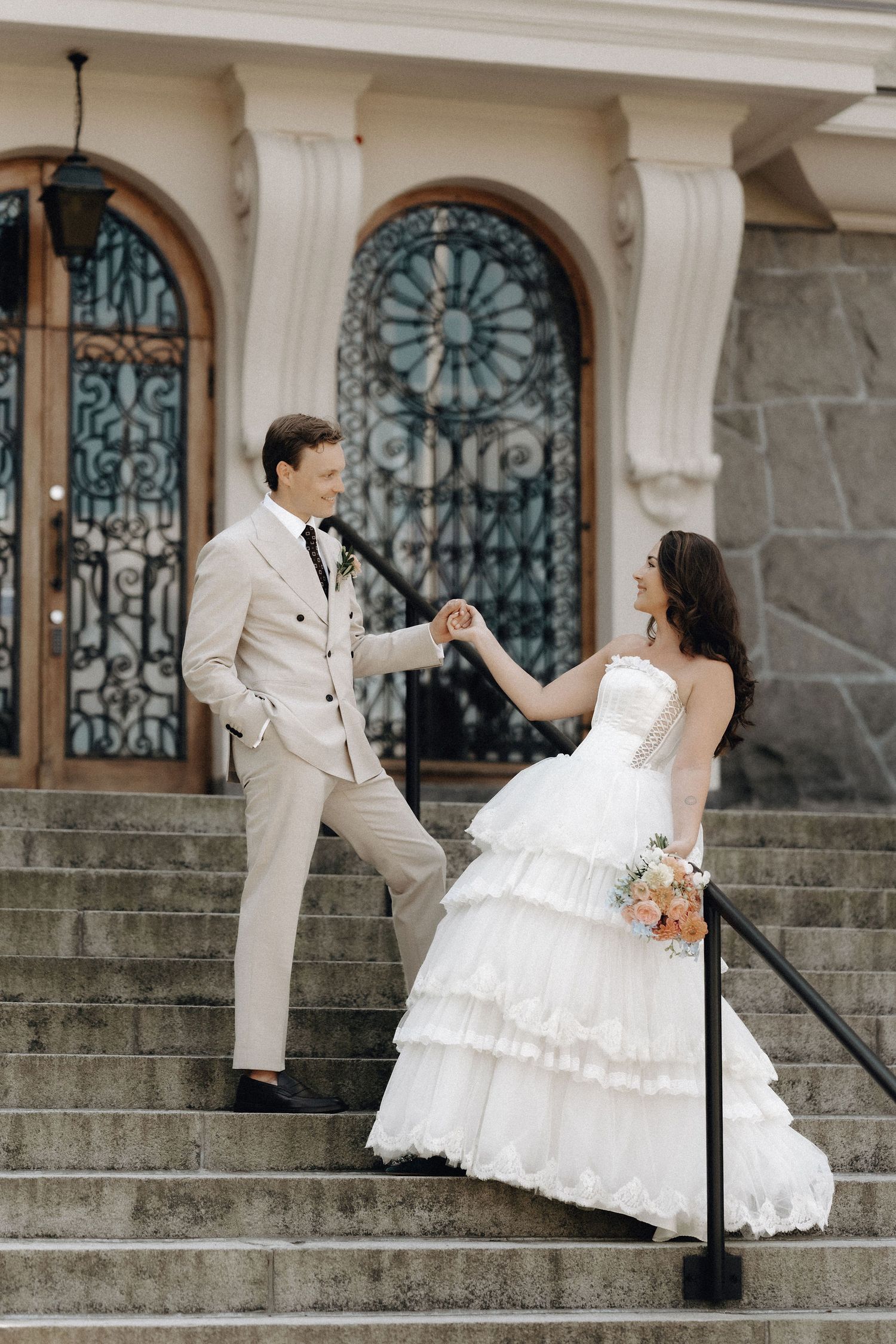 Bride and groom holding hands on grand staircase before ornate iron doors of a classic building.