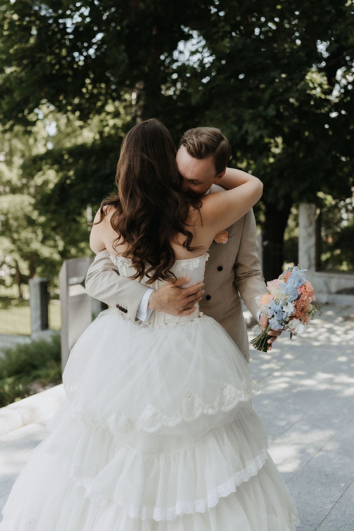 Groom in beige suit embracing and kissing bride in tiered white ballgown holding pastel bouquet in park.