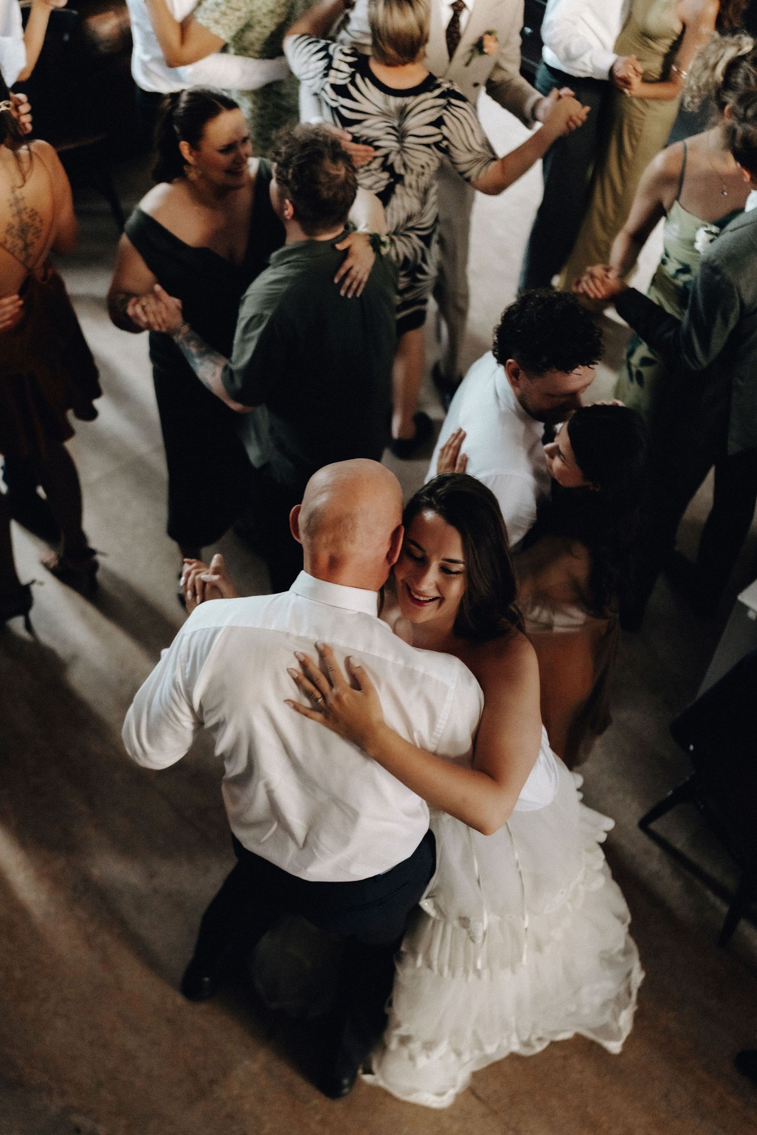 Aerial view of bride in white ruffled gown dancing joyfully with guests at wedding reception.