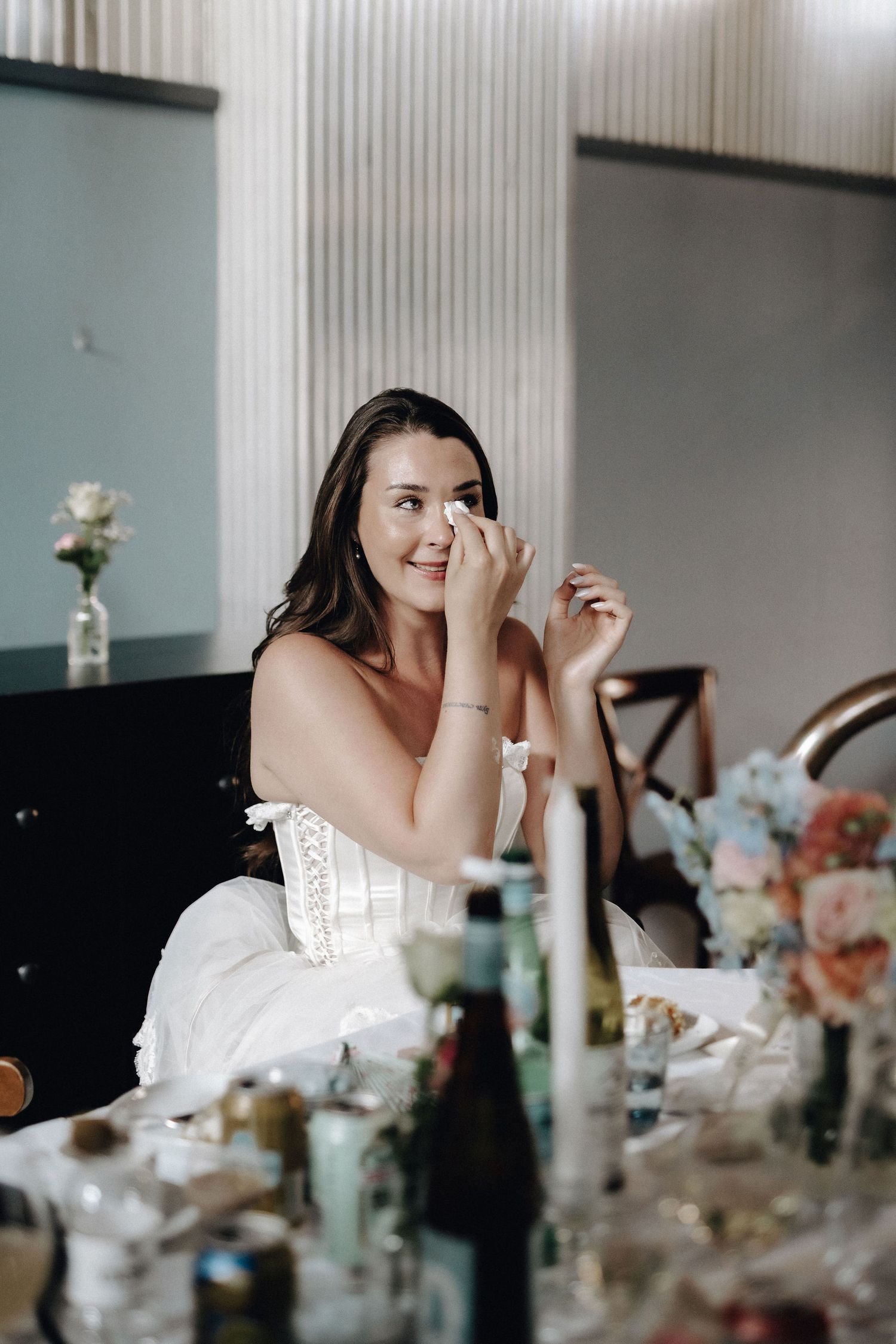 Tearful bride in white wedding dress wiping eye during emotional speech at reception.
