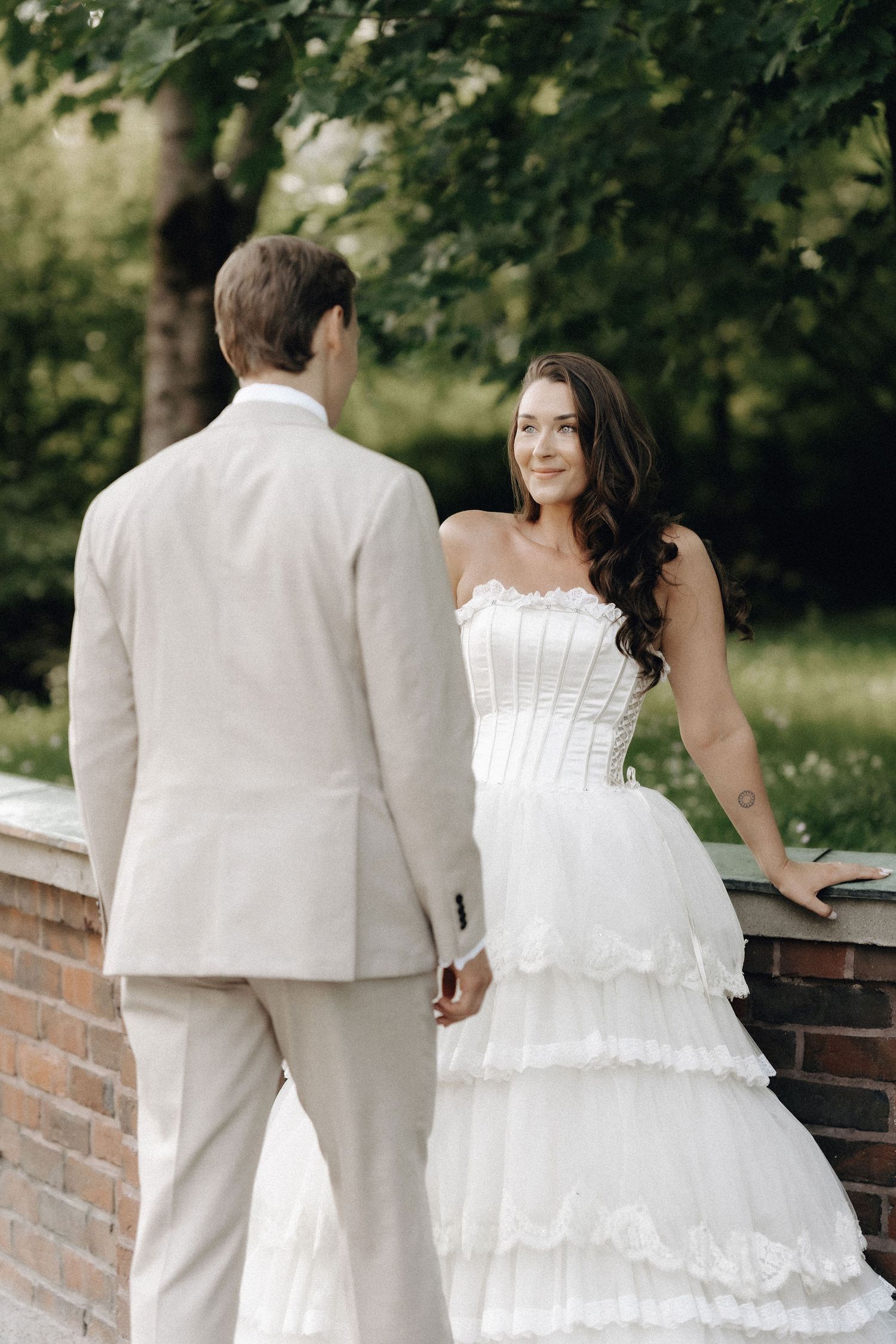 Smiling bride in strapless tiered wedding dress holding hands with groom in sunny garden.