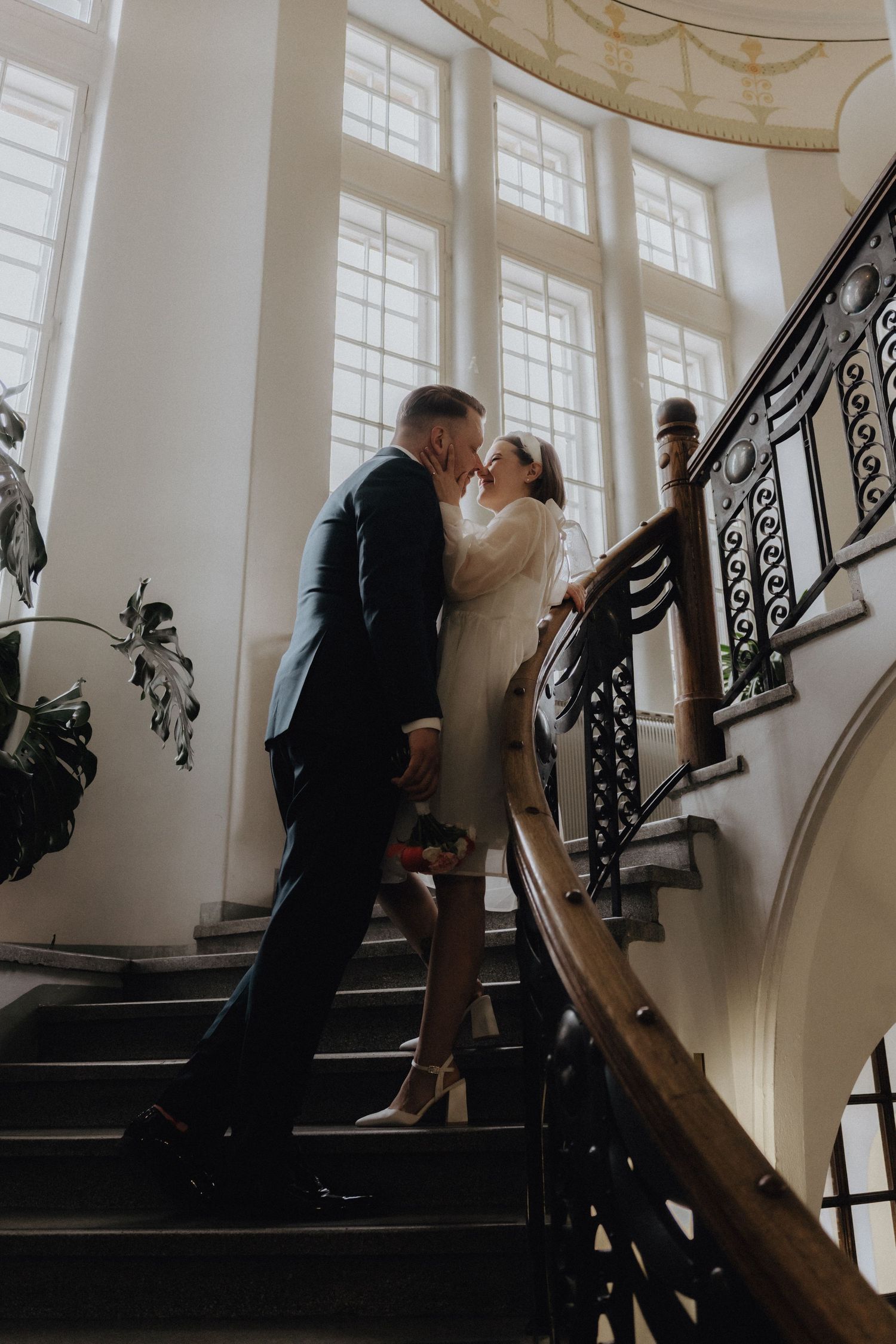 Bride and groom sharing a kiss on elegant curved staircase with ornate railing and tall windows in grand interior.