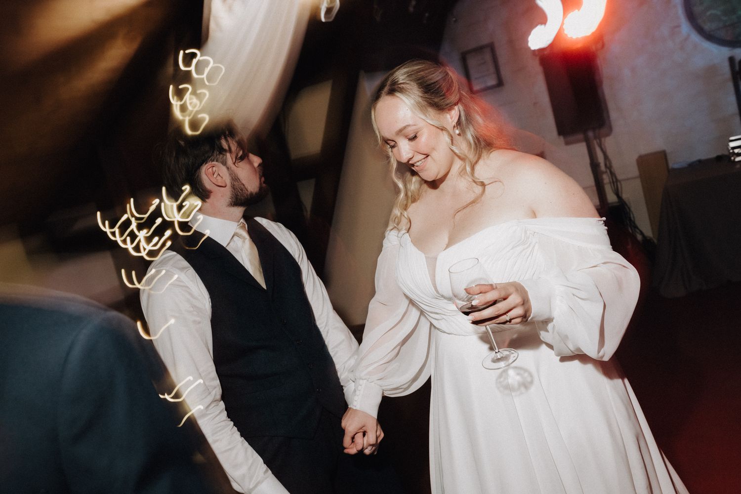 Bride in off-shoulder gown holds champagne while dancing with groom in vest under warm reception lights.