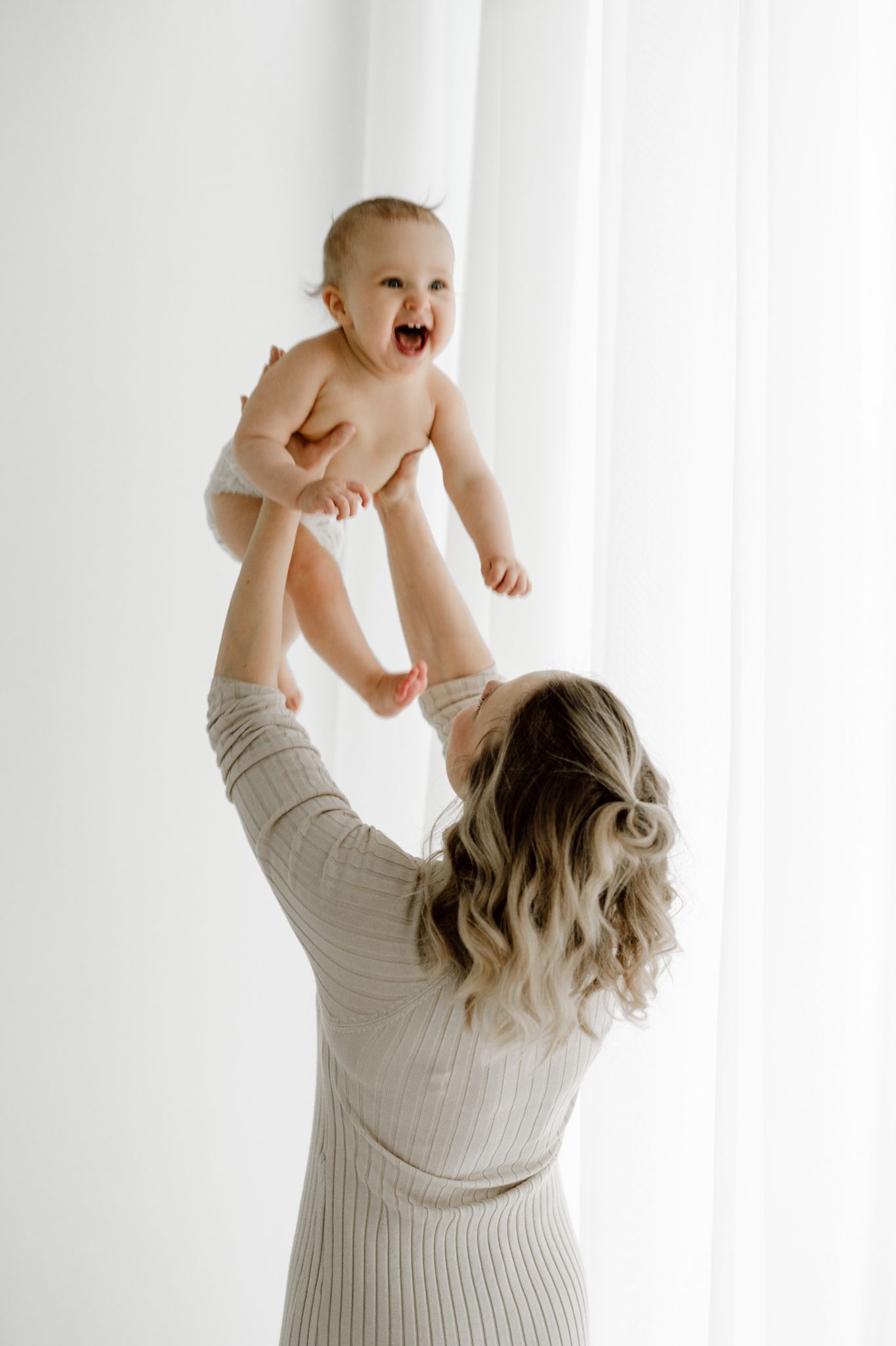 Joyful mother lifting laughing baby in diaper high in the air against white curtains.