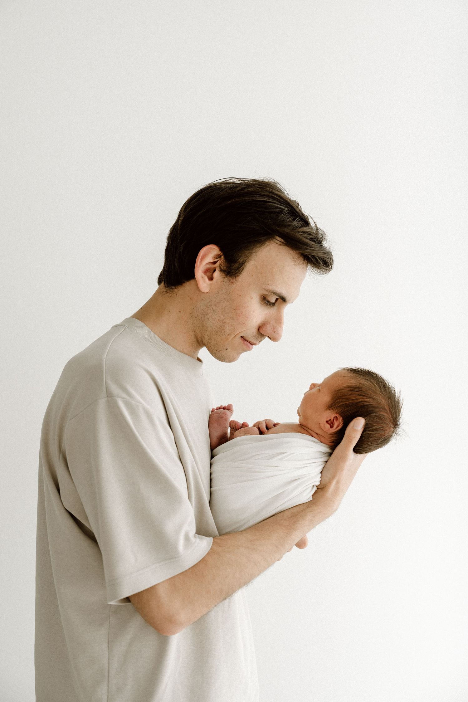 Dark-haired father in beige shirt gently cradling swaddled newborn baby against white background.