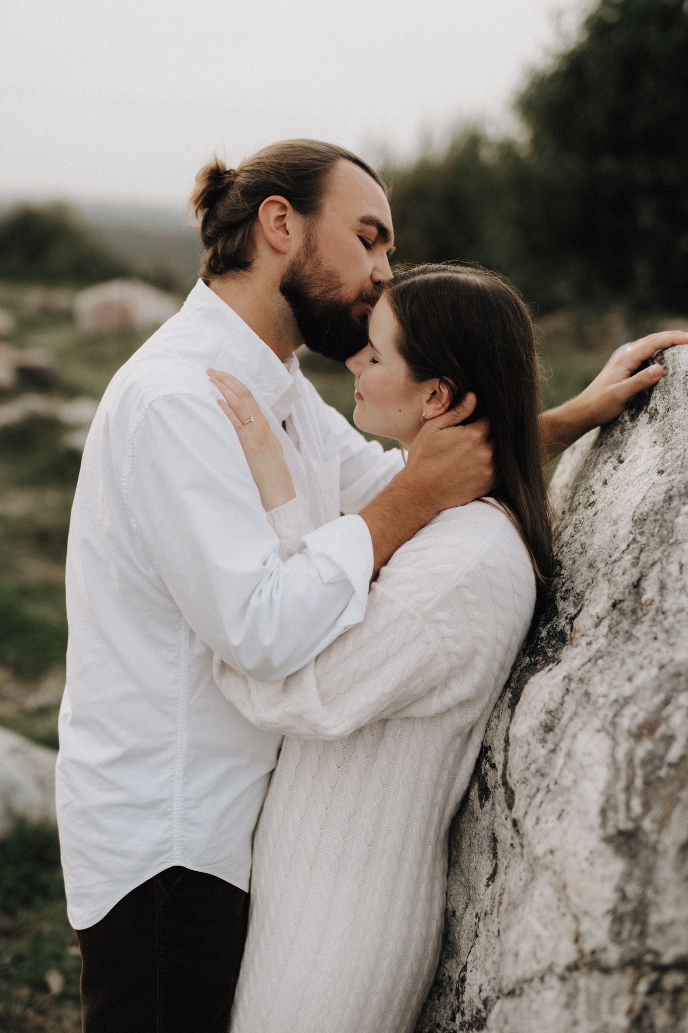 Bearded man kissing woman leaning against a rock, both dressed in white in a natural setting.
