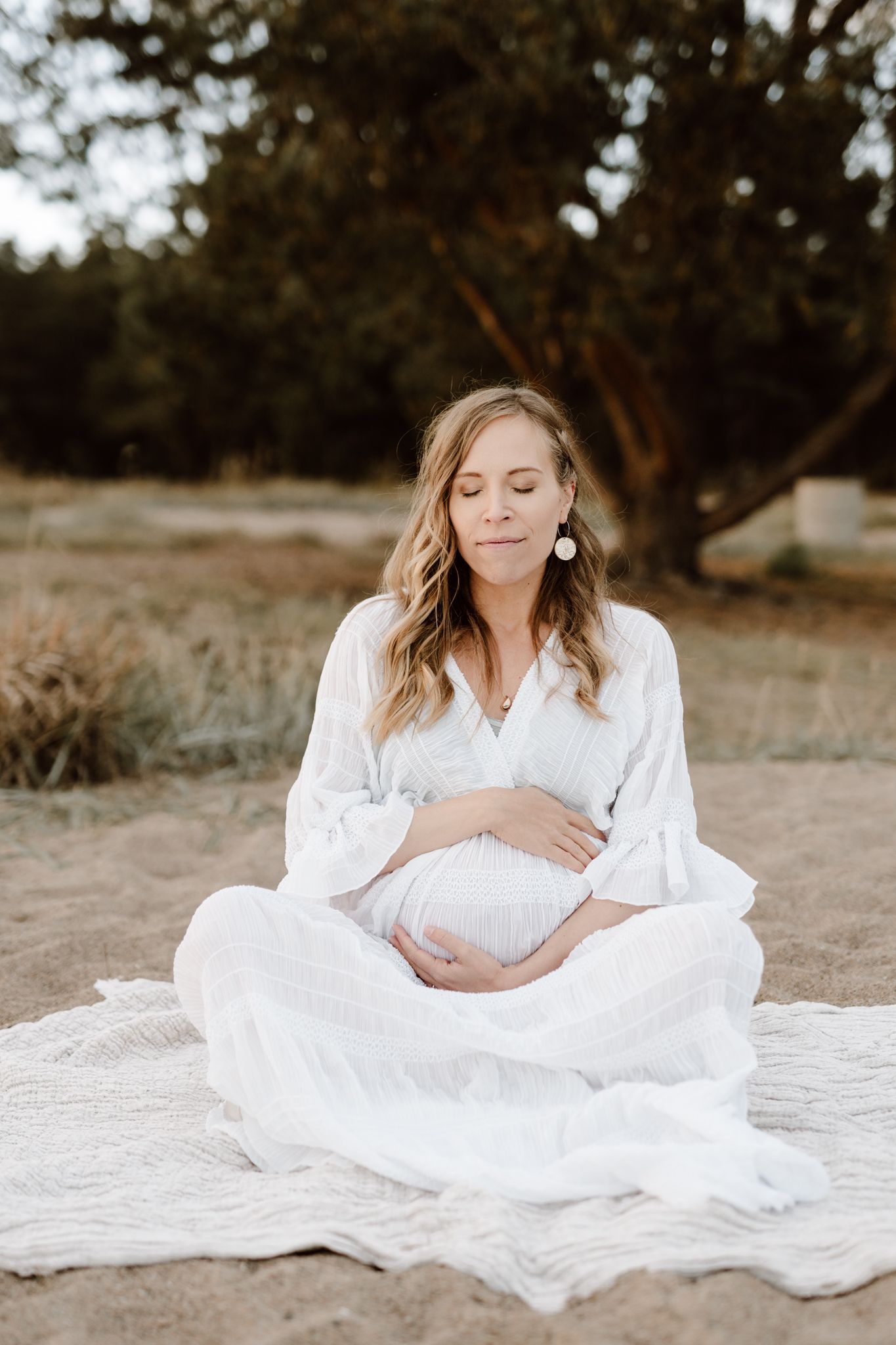 Pregnant woman in white dress sitting cross-legged on blanket outdoors with eyes closed.