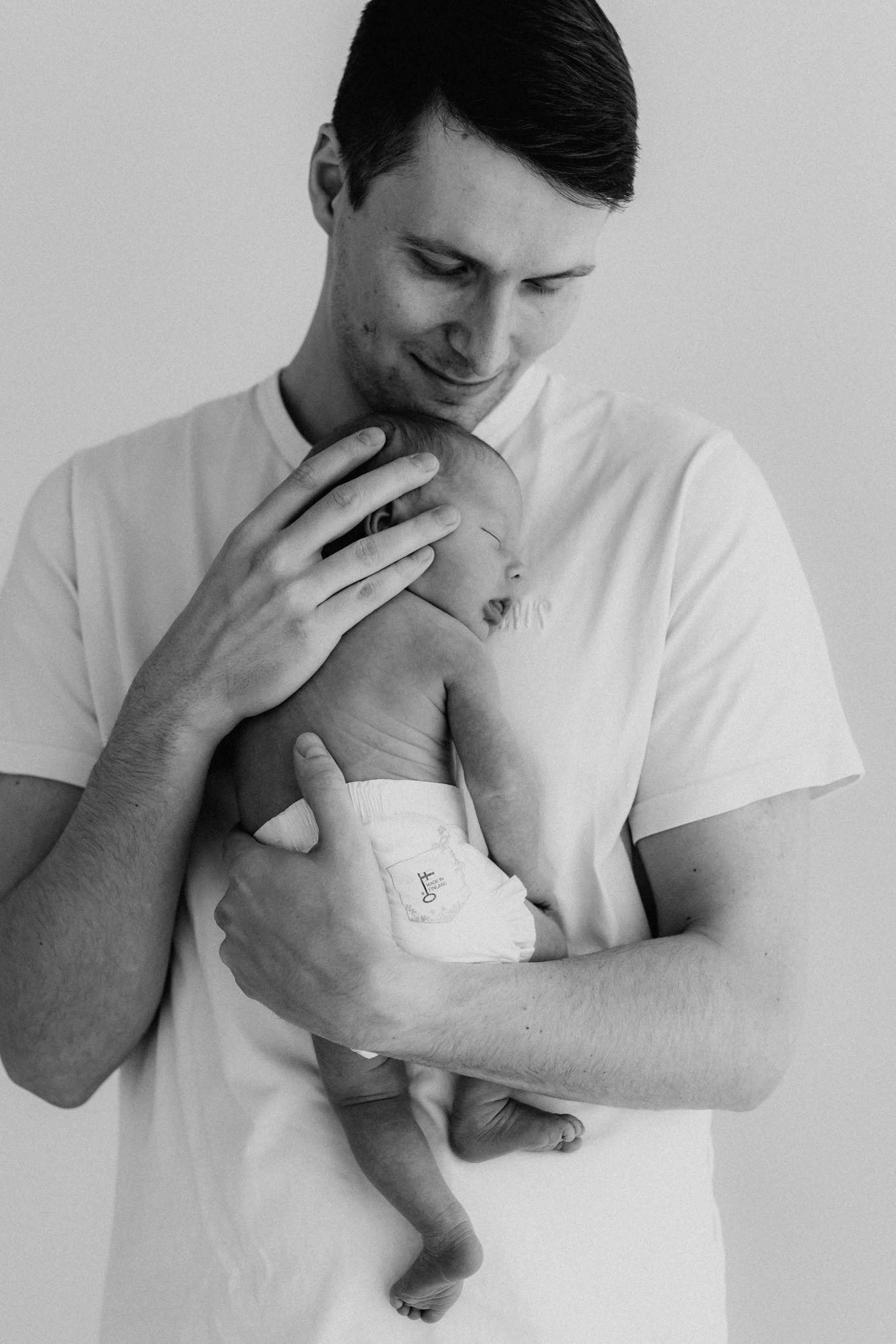 Black and white photo of a smiling father in white t-shirt carefully holding his newborn baby.