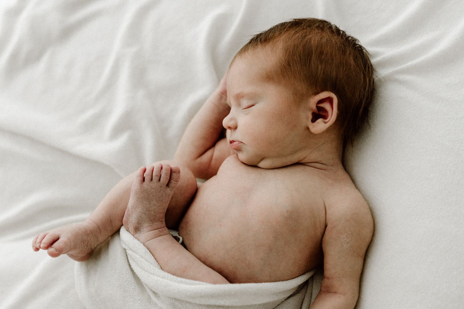 Side view of sleeping newborn baby wrapped in white swaddle on white blanket.