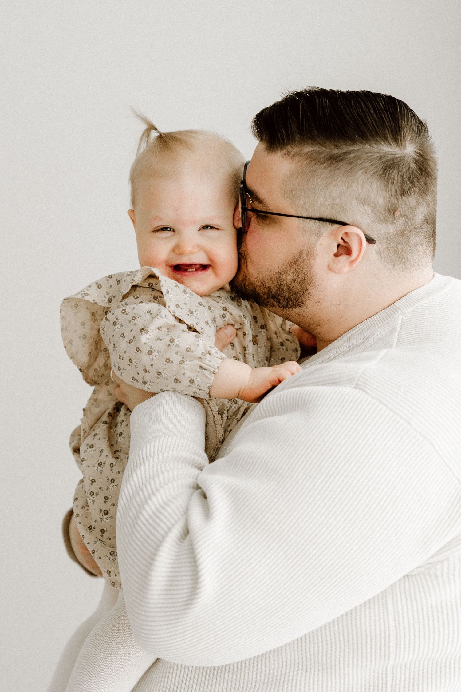 A bearded father in glasses kisses his laughing baby girl in a floral outfit against a white background.