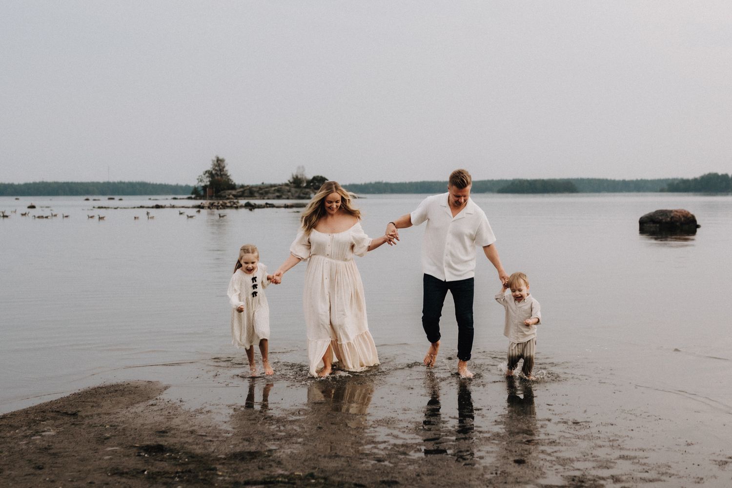 Happy family of four holding hands and splashing through shallow water on a serene lake beach.