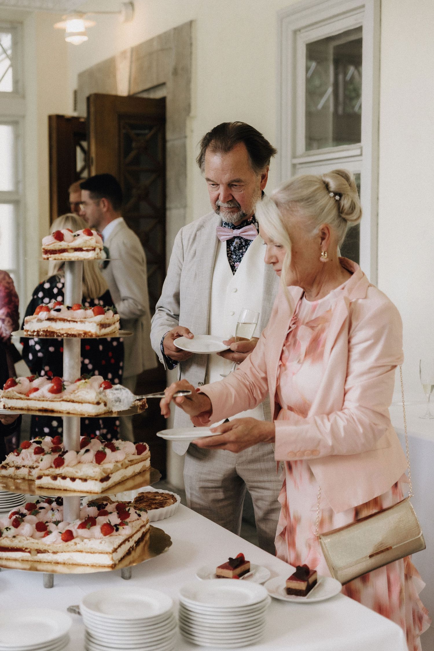 Older couple serving themselves slices of a tiered strawberry wedding cake at the reception table.