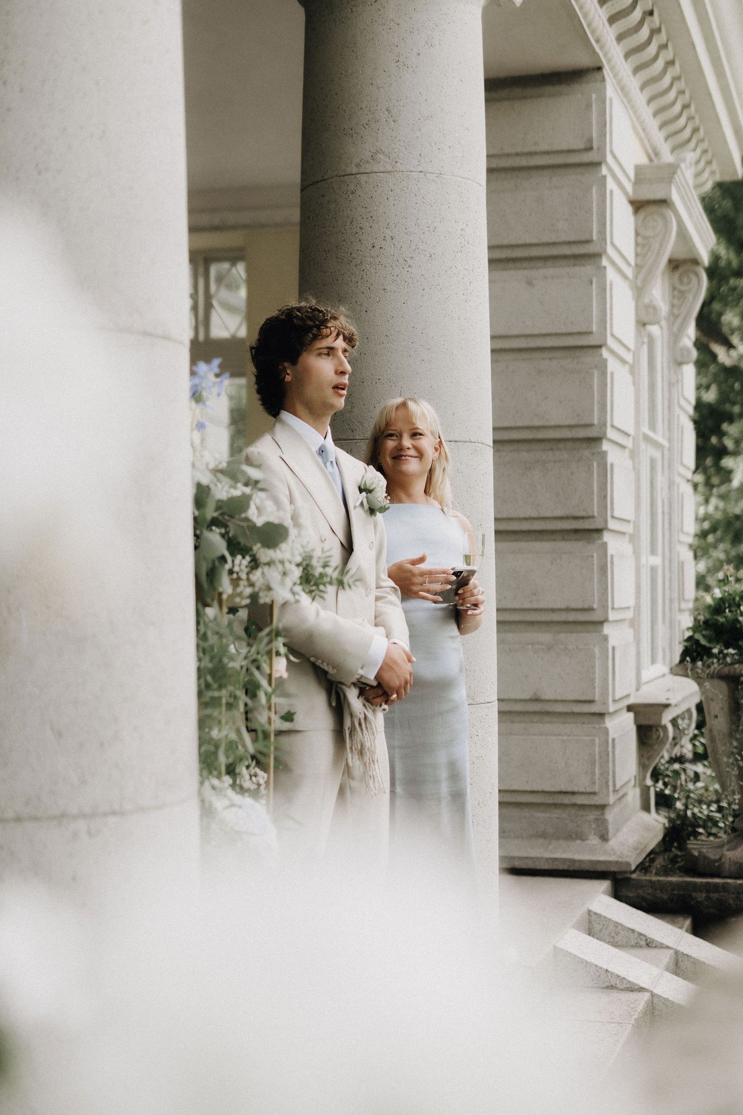 Groom in beige suit stands with an older woman beside stone columns at a wedding venue.