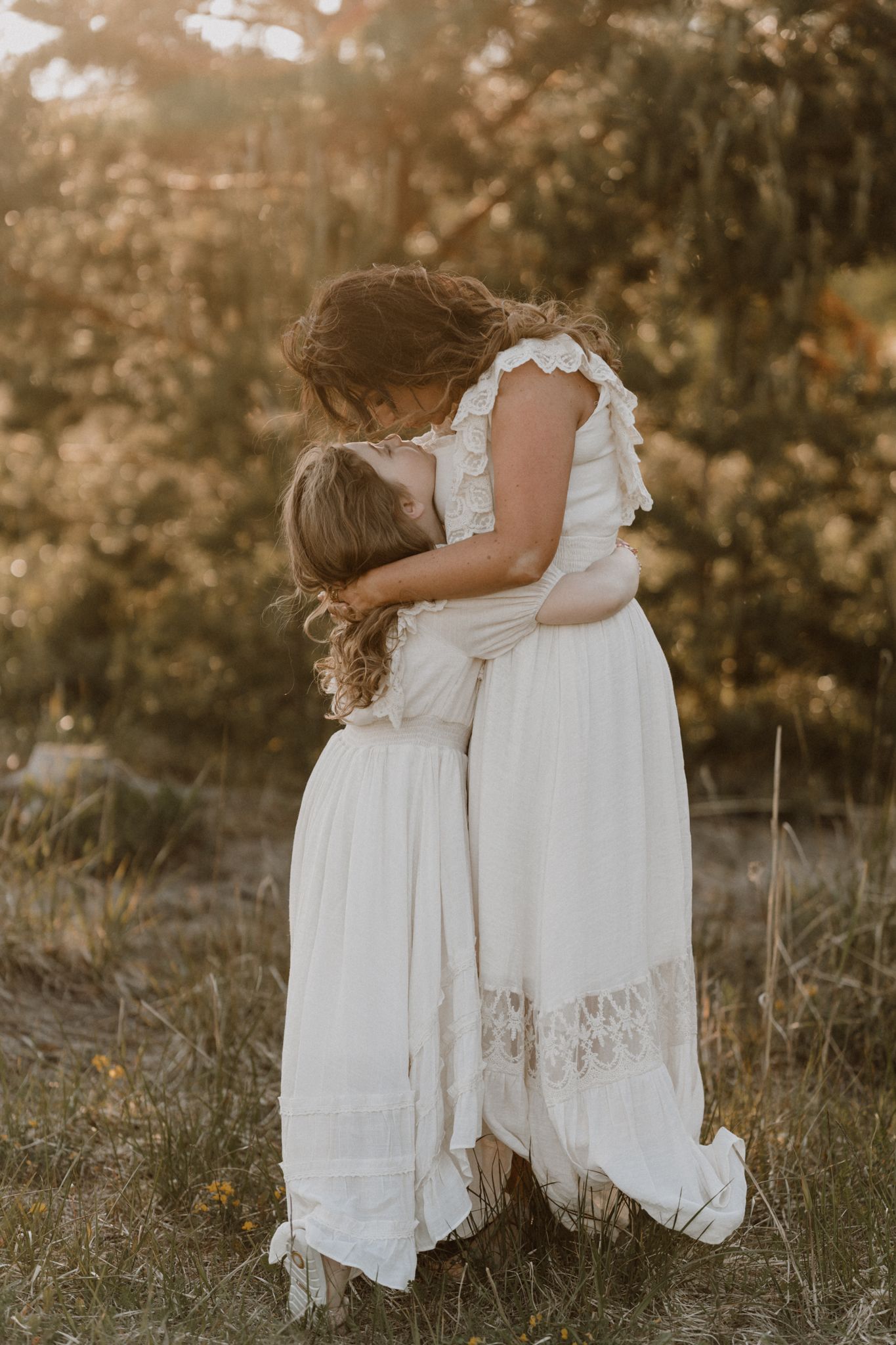 A mother and young daughter share a warm embrace in white dresses among golden sunlit trees.