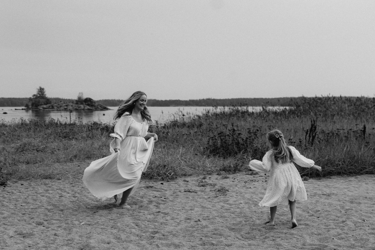 A mother and daughter in white dresses run freely along a sandy beach in a black and white photo.