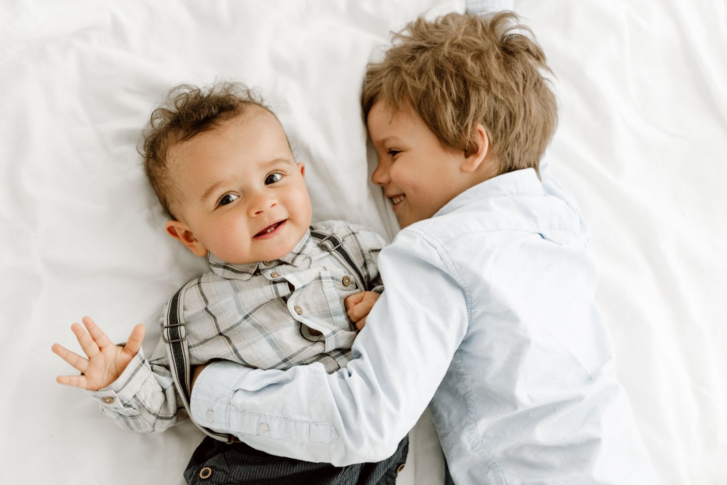Two young brothers lying on white bedding, older boy smiling at happy baby sibling.