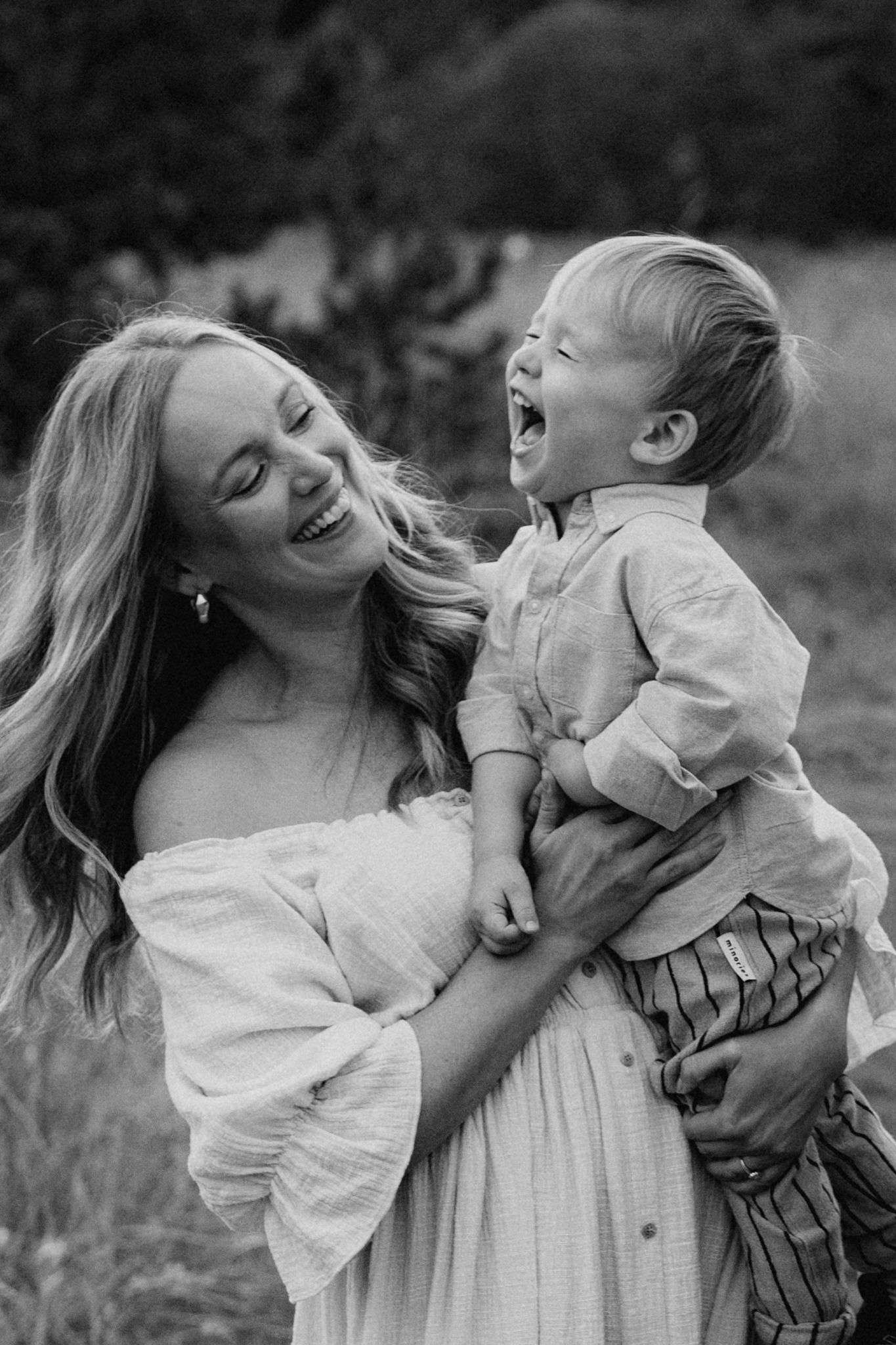Black and white photo of laughing mother holding giggling toddler boy outdoors.