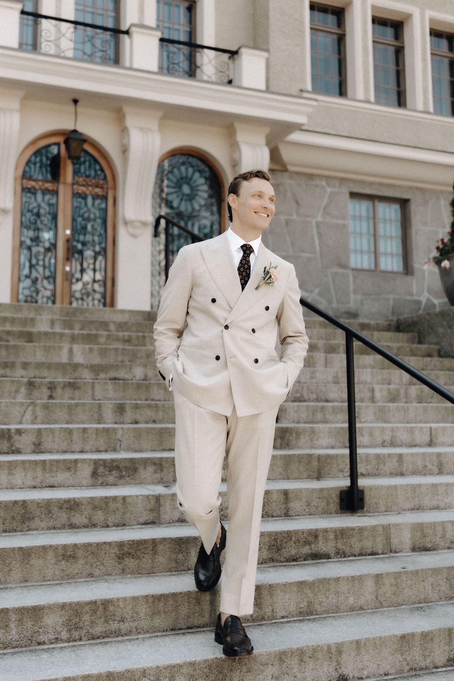 Handsome groom in cream double-breasted suit with boutonniere posing on steps of an elegant venue.