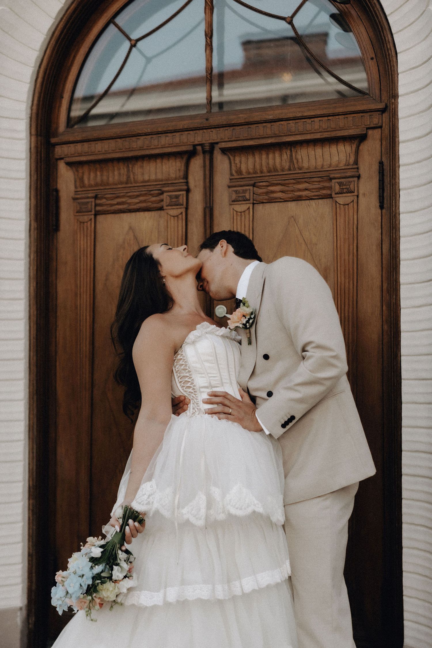 Bride and groom sharing a romantic kiss in front of ornate wooden doors on their wedding day.