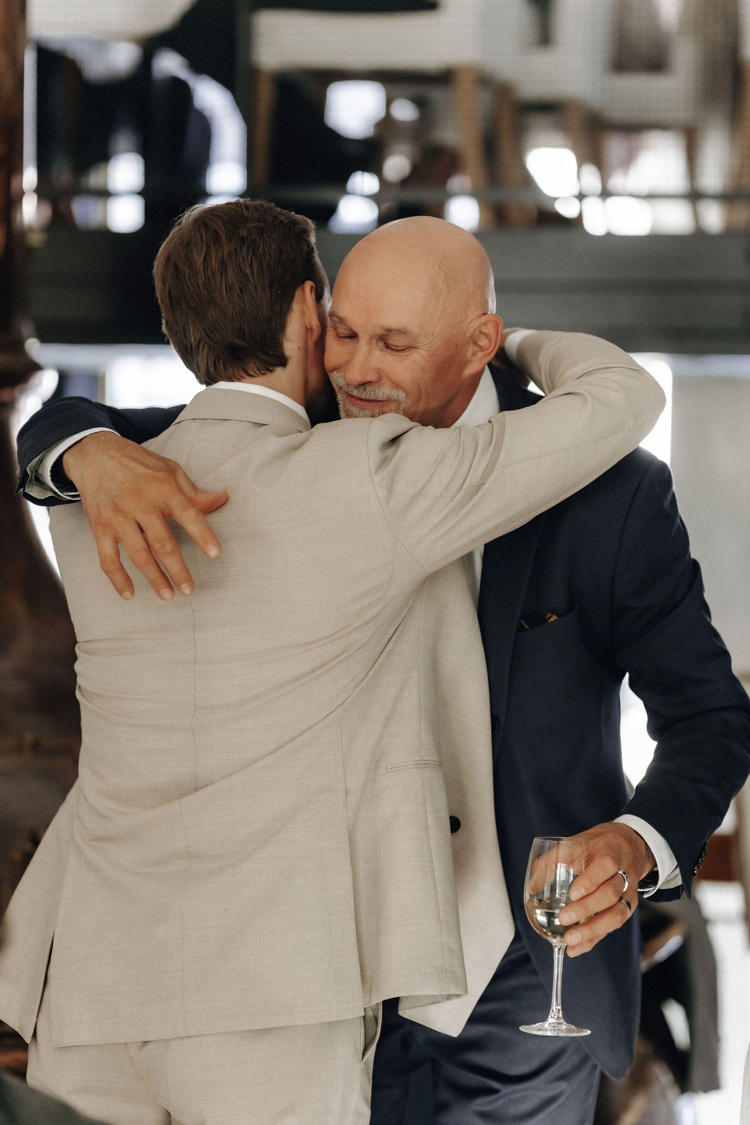 Two men in suits sharing an emotional hug at a wedding reception with champagne glass.
