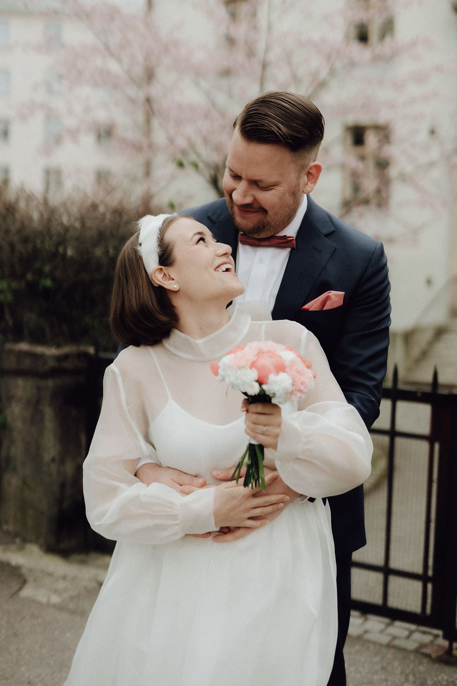 Groom in navy suit embracing laughing bride in vintage white dress with headband, holding pink bouquet near cherry blossoms.