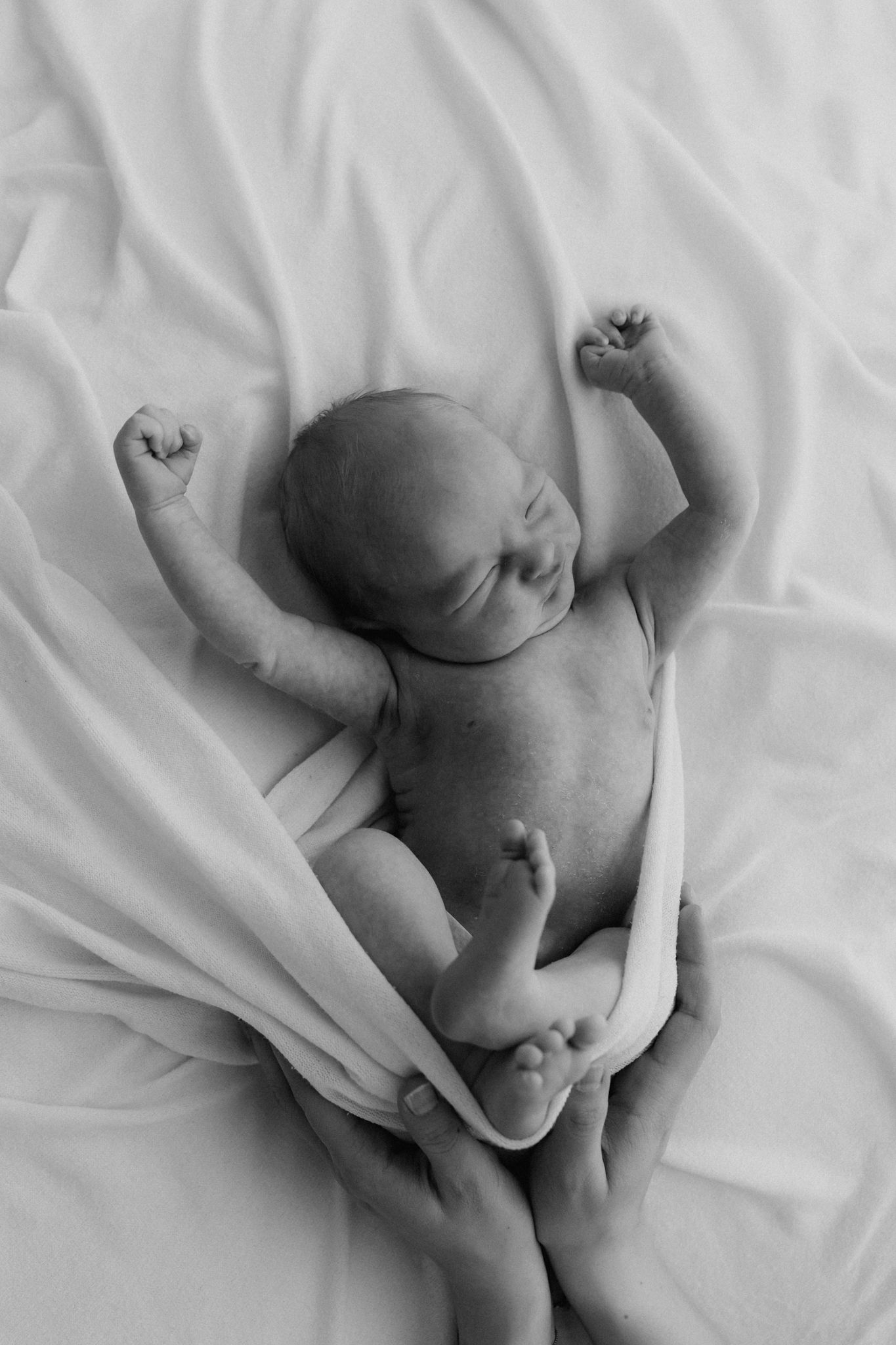Black and white overhead photo of newborn baby stretching arms while lying on white fabric.