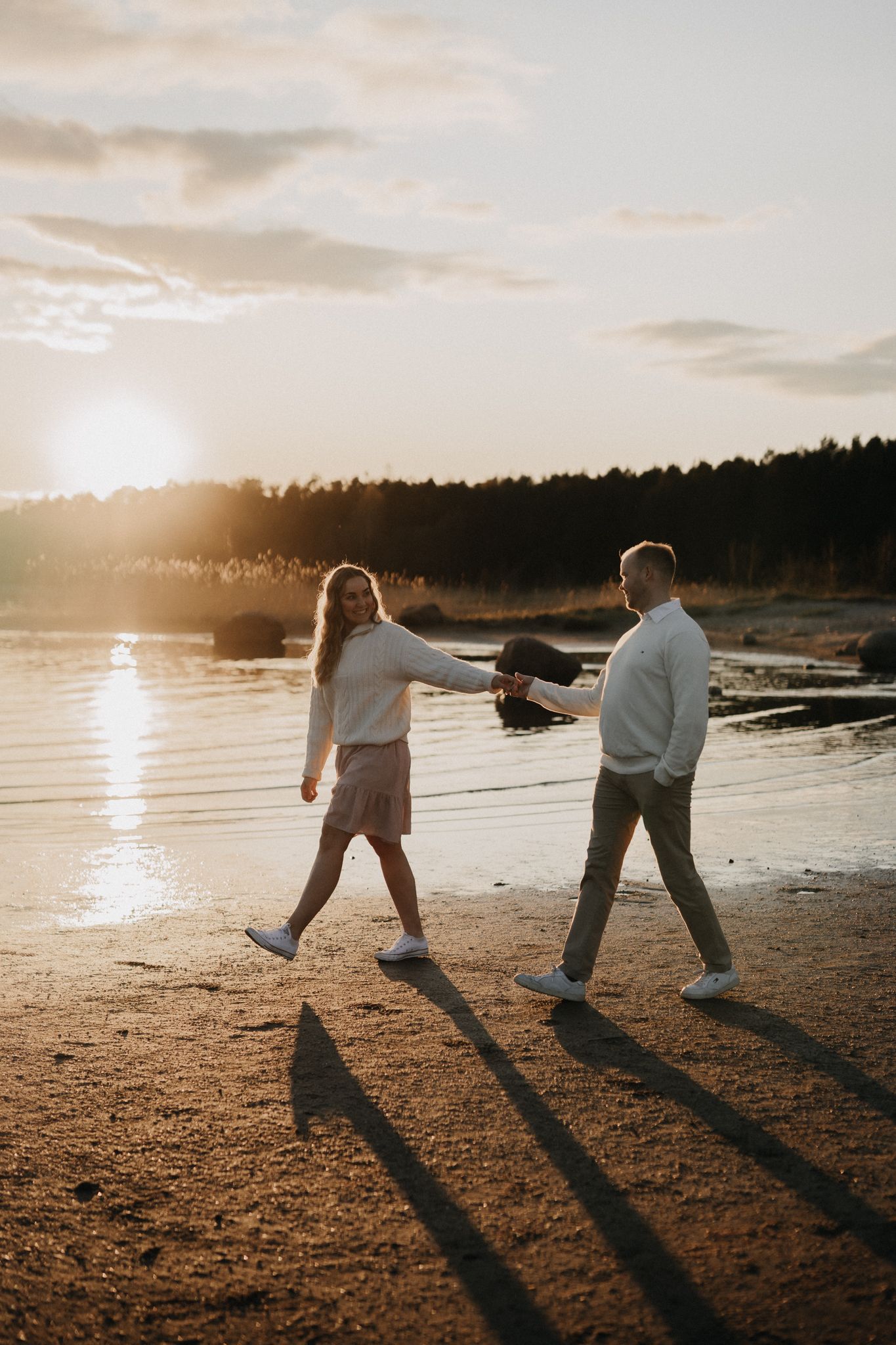 Couple walks hand-in-hand along a sandy beach at sunset, casting long shadows in warm golden light.