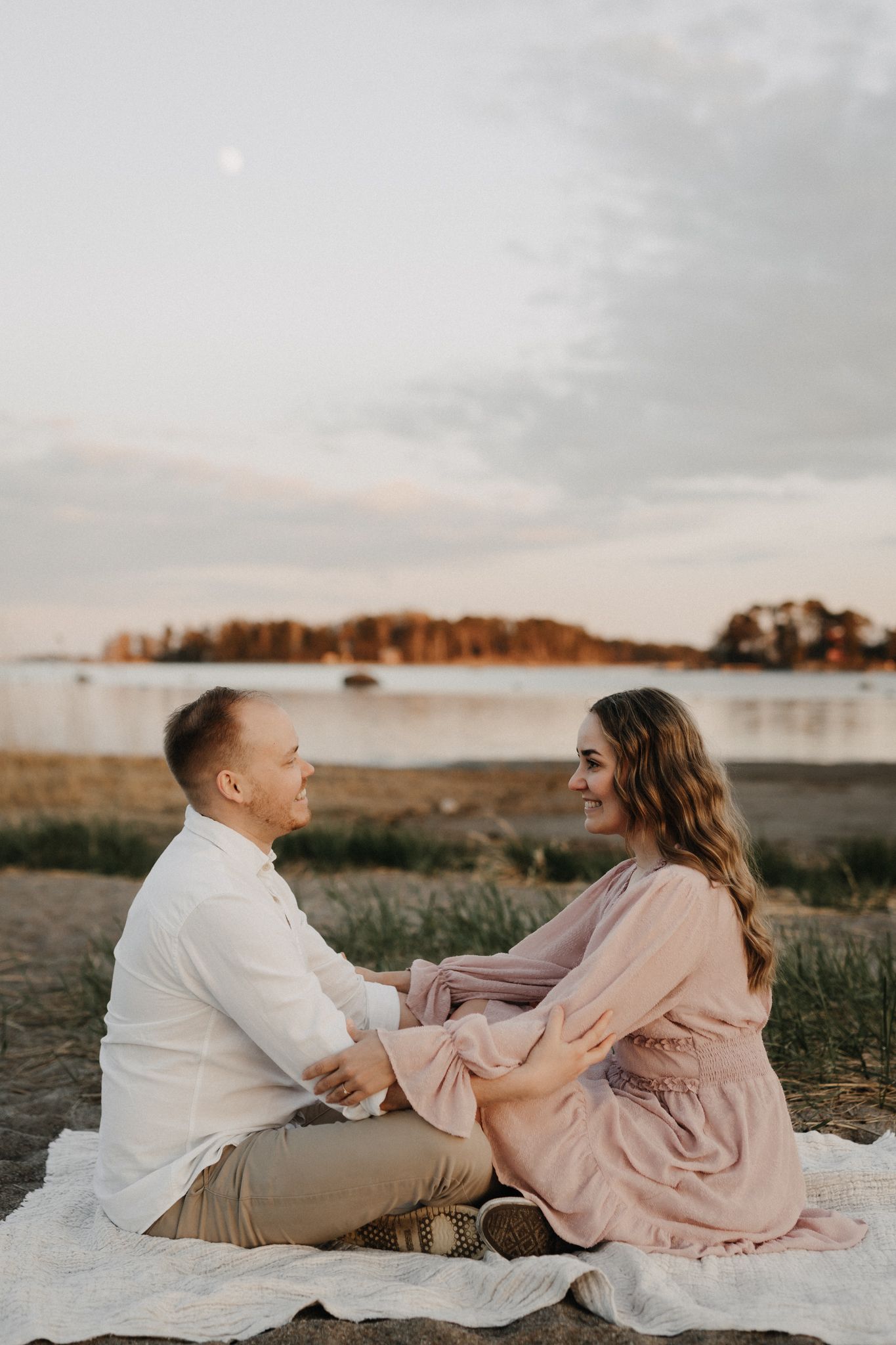 Couple sitting face-to-face on blanket by lake at dusk, holding hands and smiling warmly.