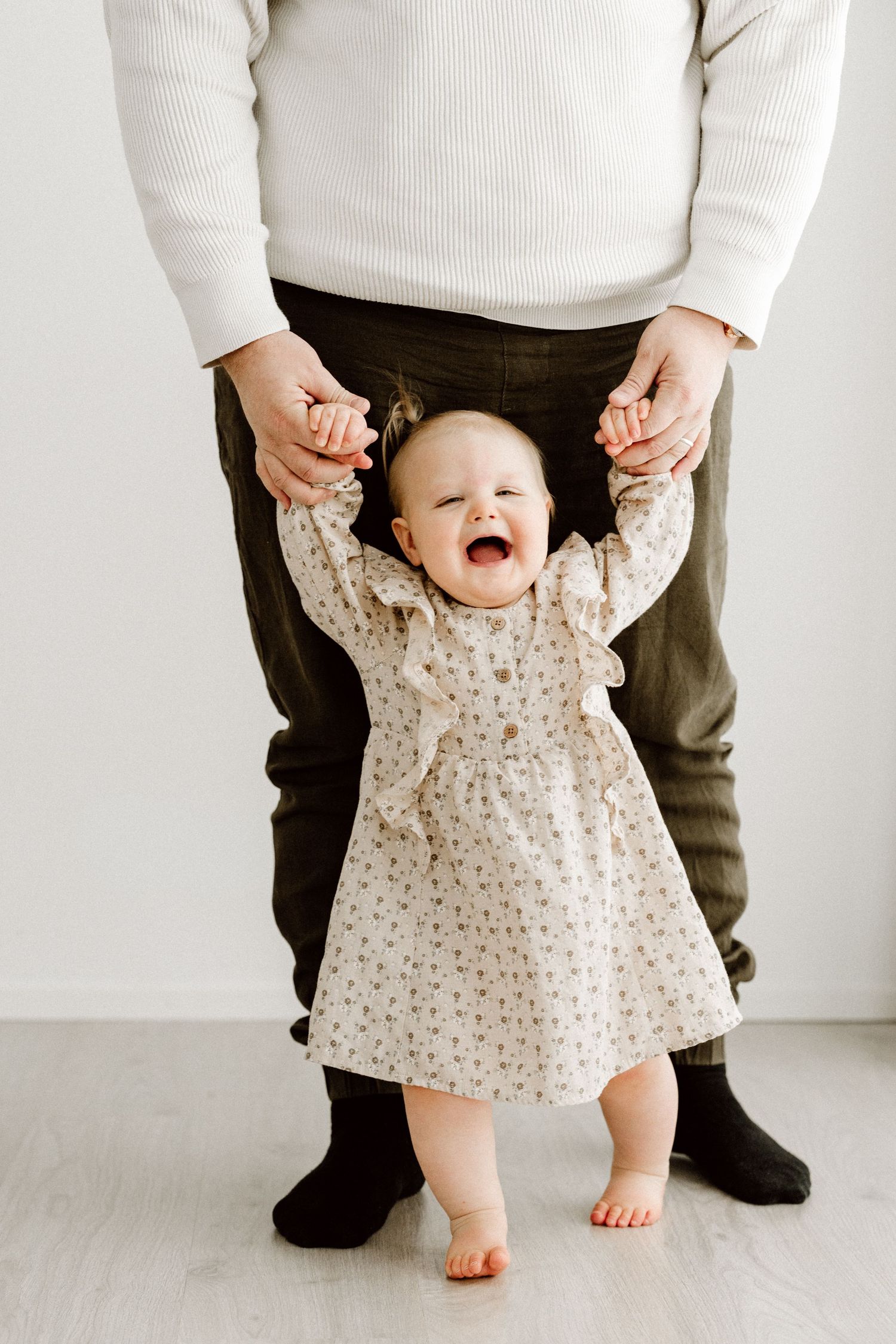 A laughing baby girl in a floral dress stands barefoot while being supported by her parent's hands.