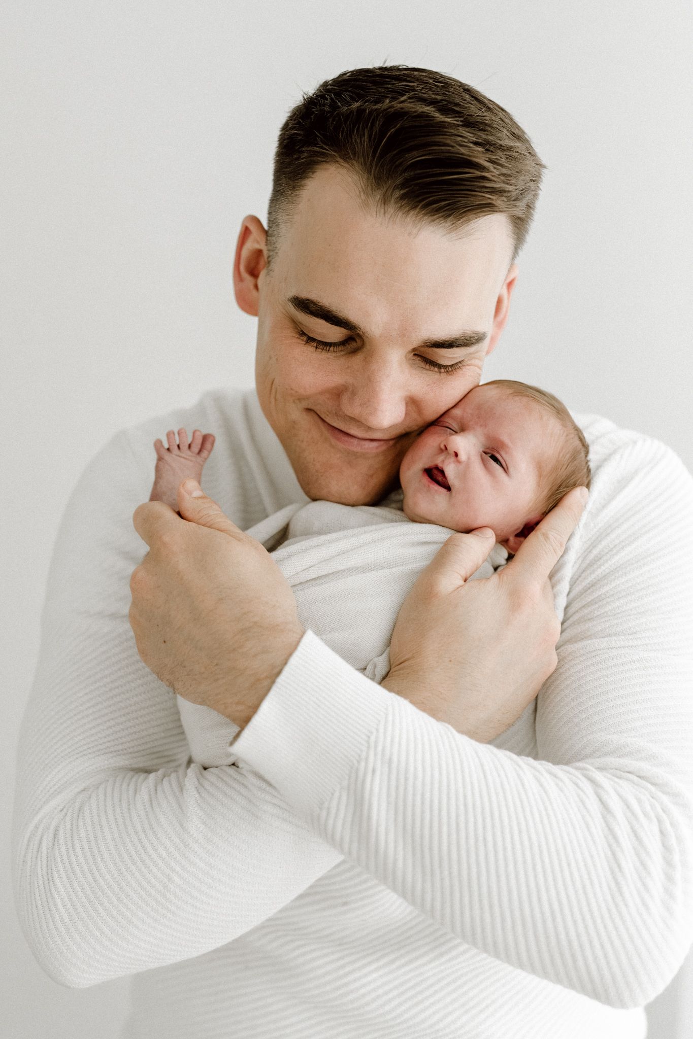 Loving father in white sweater gently cradles smiling newborn baby against his cheek in a studio portrait.