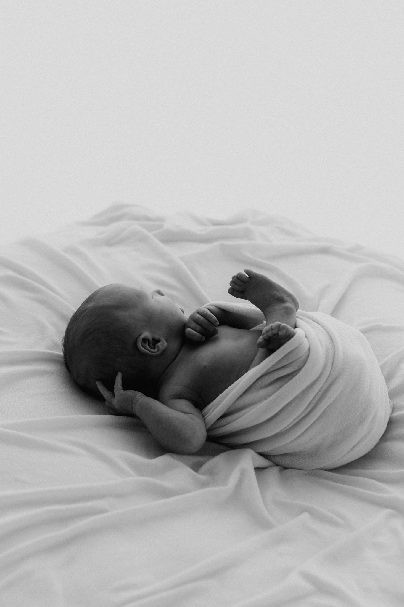 Black and white photo of an alert newborn baby loosely wrapped in white swaddle on bedding.
