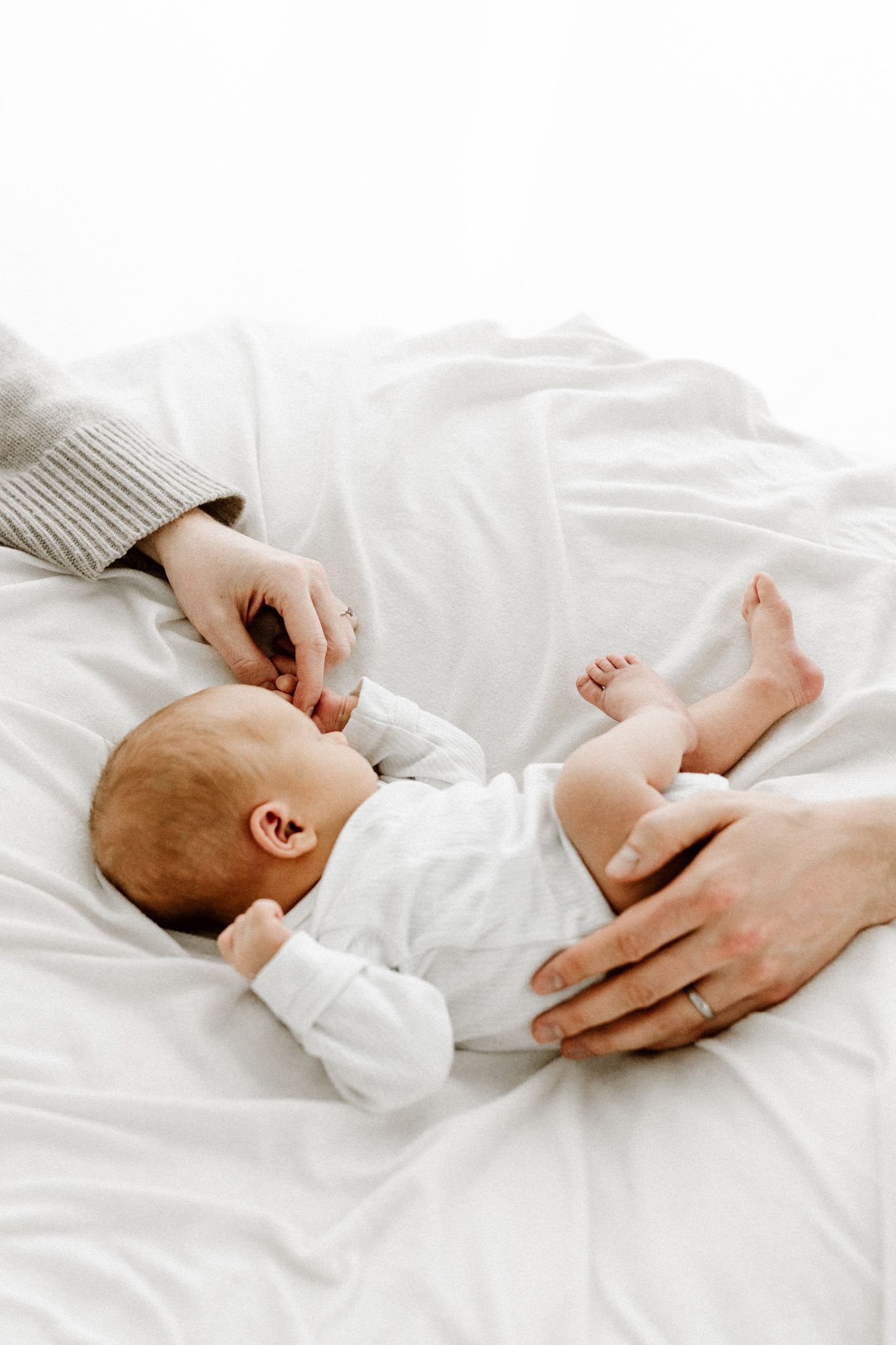 Parent hands gently cradling a newborn baby in white outfit on soft white bedding.