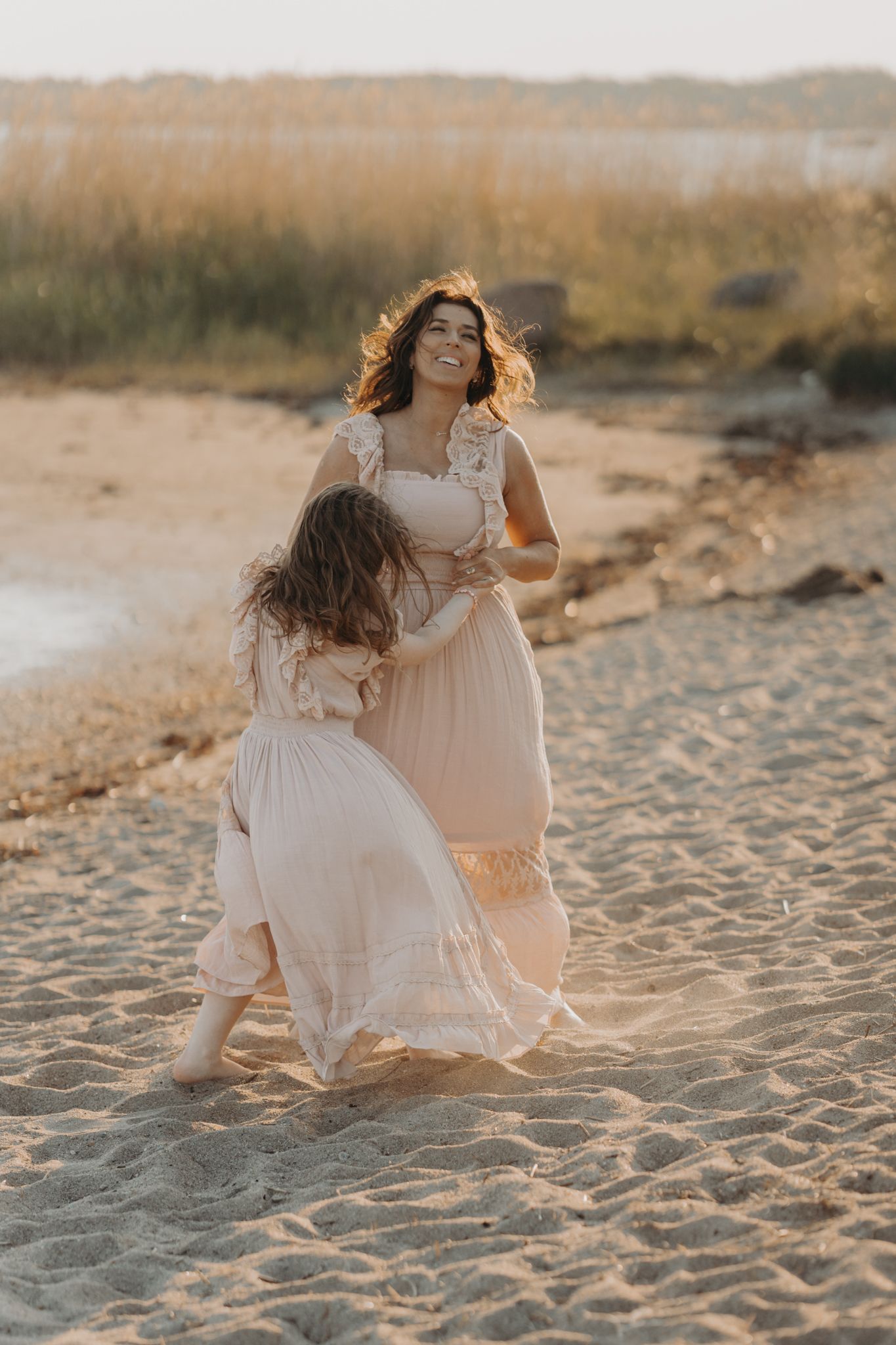 A smiling mother in a blush dress twirls joyfully with her young daughter on a sunlit sandy beach.
