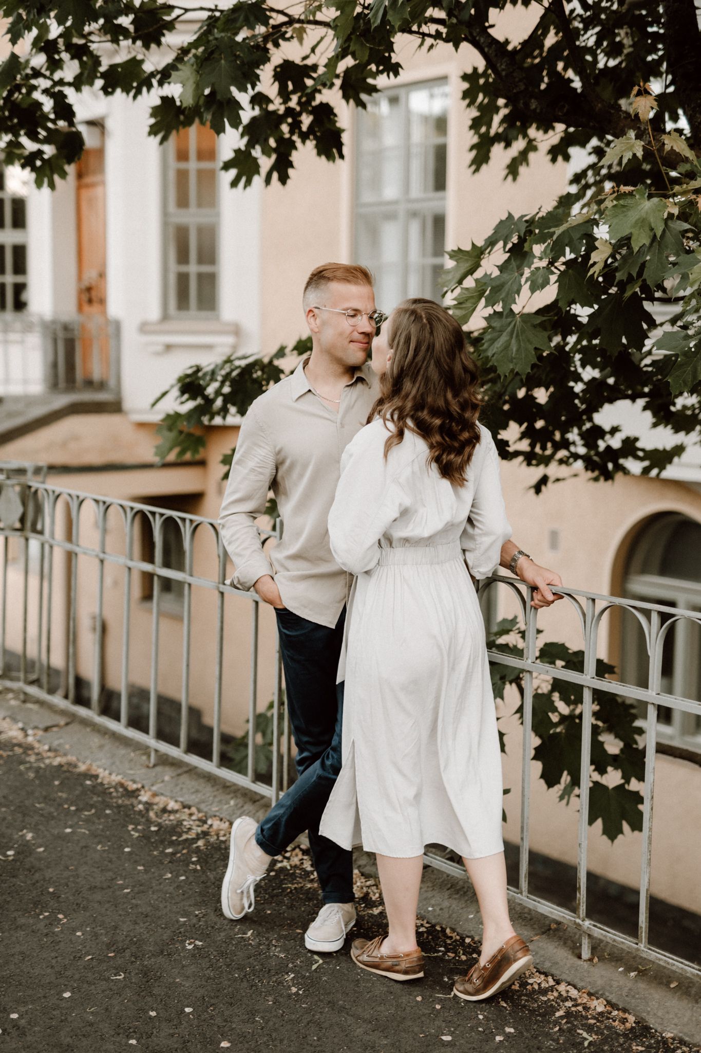 Couple in coordinating beige outfits sharing a kiss by an iron fence with lush green trees.
