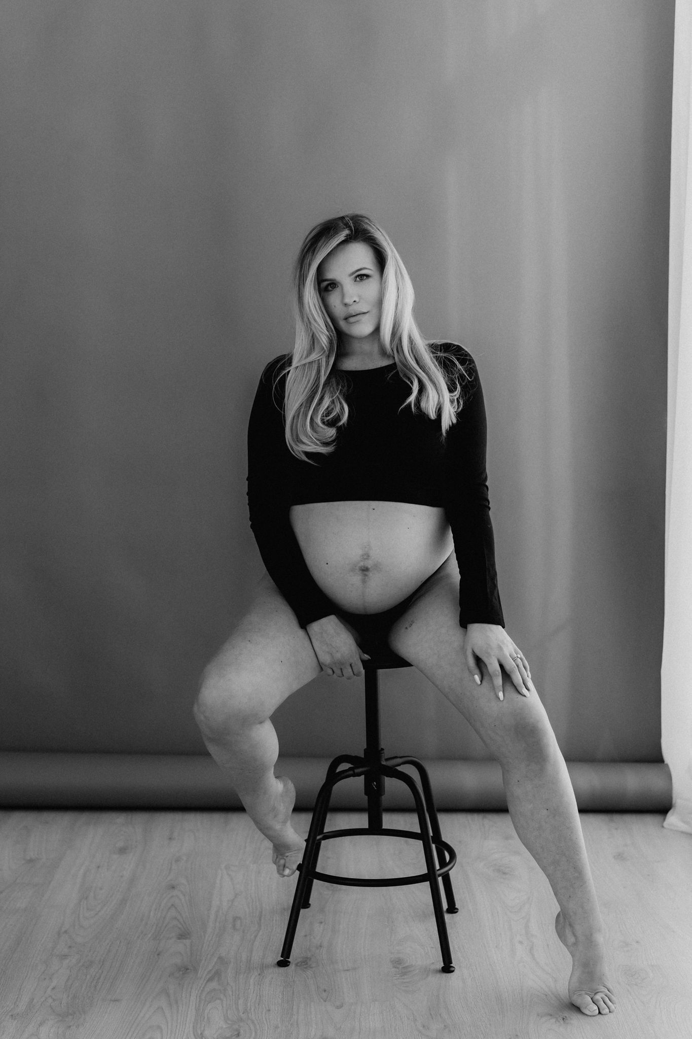 Pregnant blonde woman in black outfit seated on stool in black and white studio portrait.