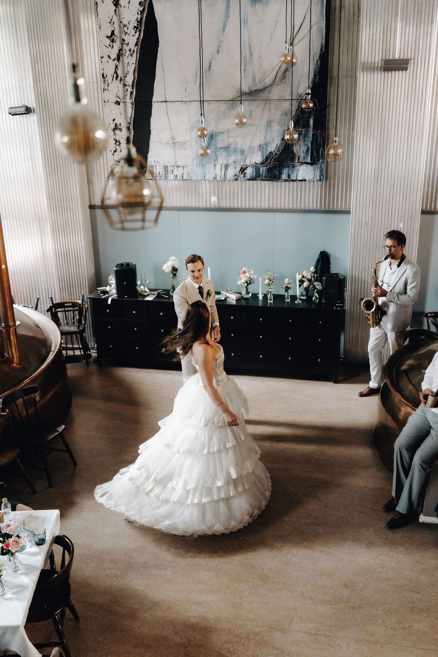 Bride in tiered white gown twirls during first dance as saxophonist performs at elegant indoor wedding reception.