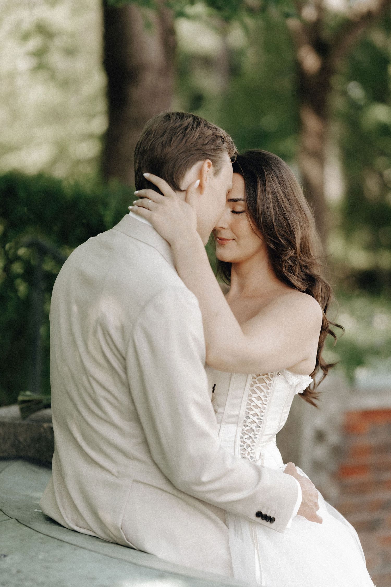 Newlyweds share an intimate almost-kiss while seated together in a sunlit outdoor garden setting.