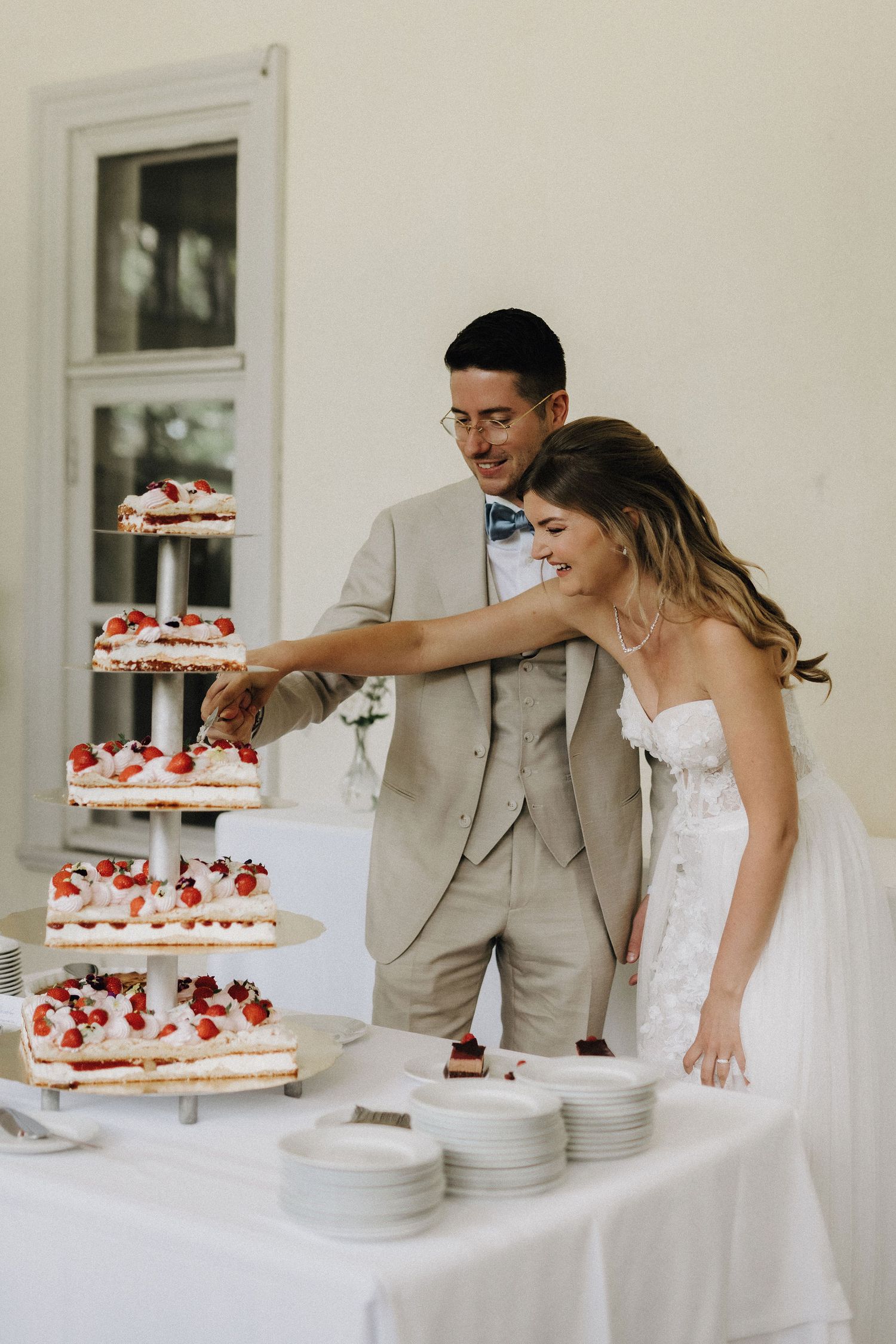 Smiling bride and groom in cream attire cutting a beautiful multi-tiered strawberry wedding cake together.