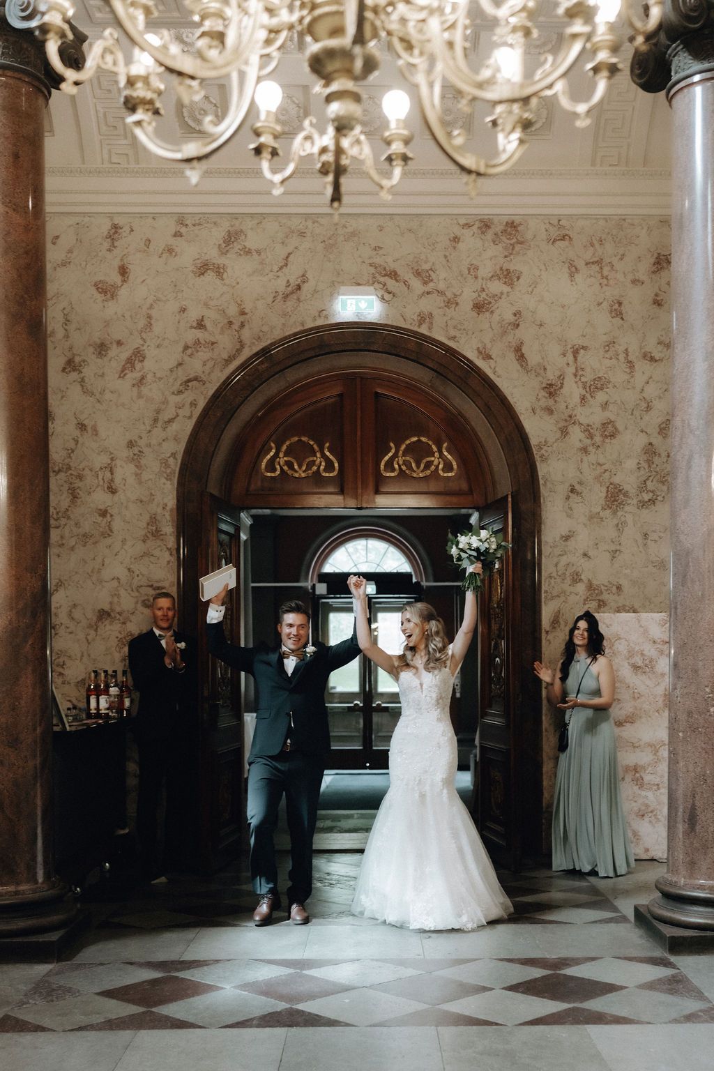 Joyful newlyweds raise hands in celebration under chandelier in elegant marble wedding venue.