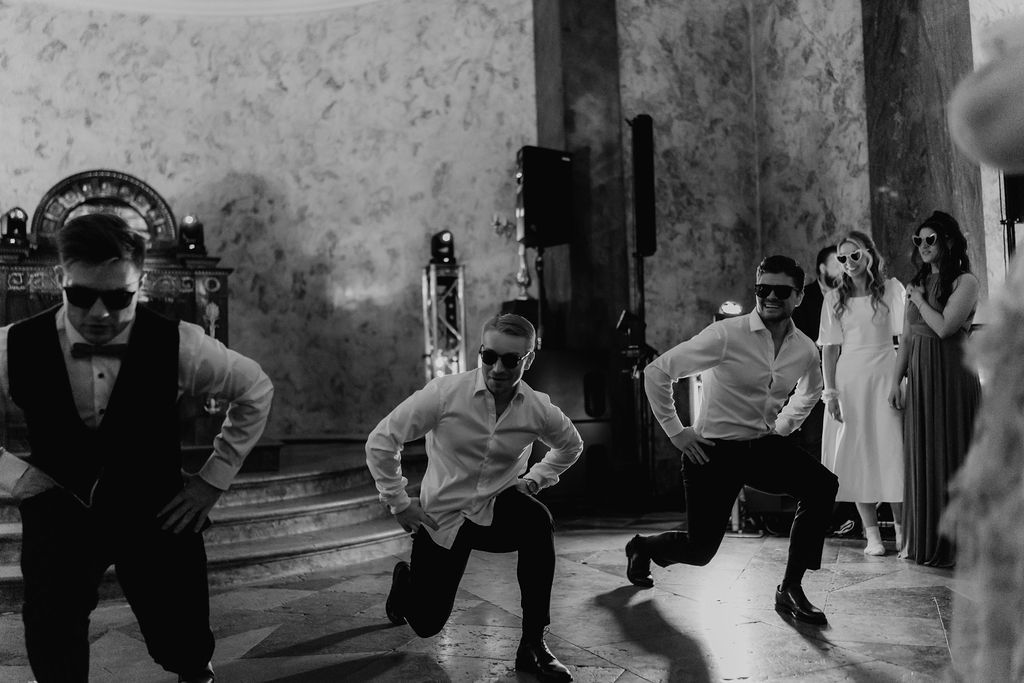 Groomsmen perform dramatic synchronized squat dance move in black and white wedding reception photo.