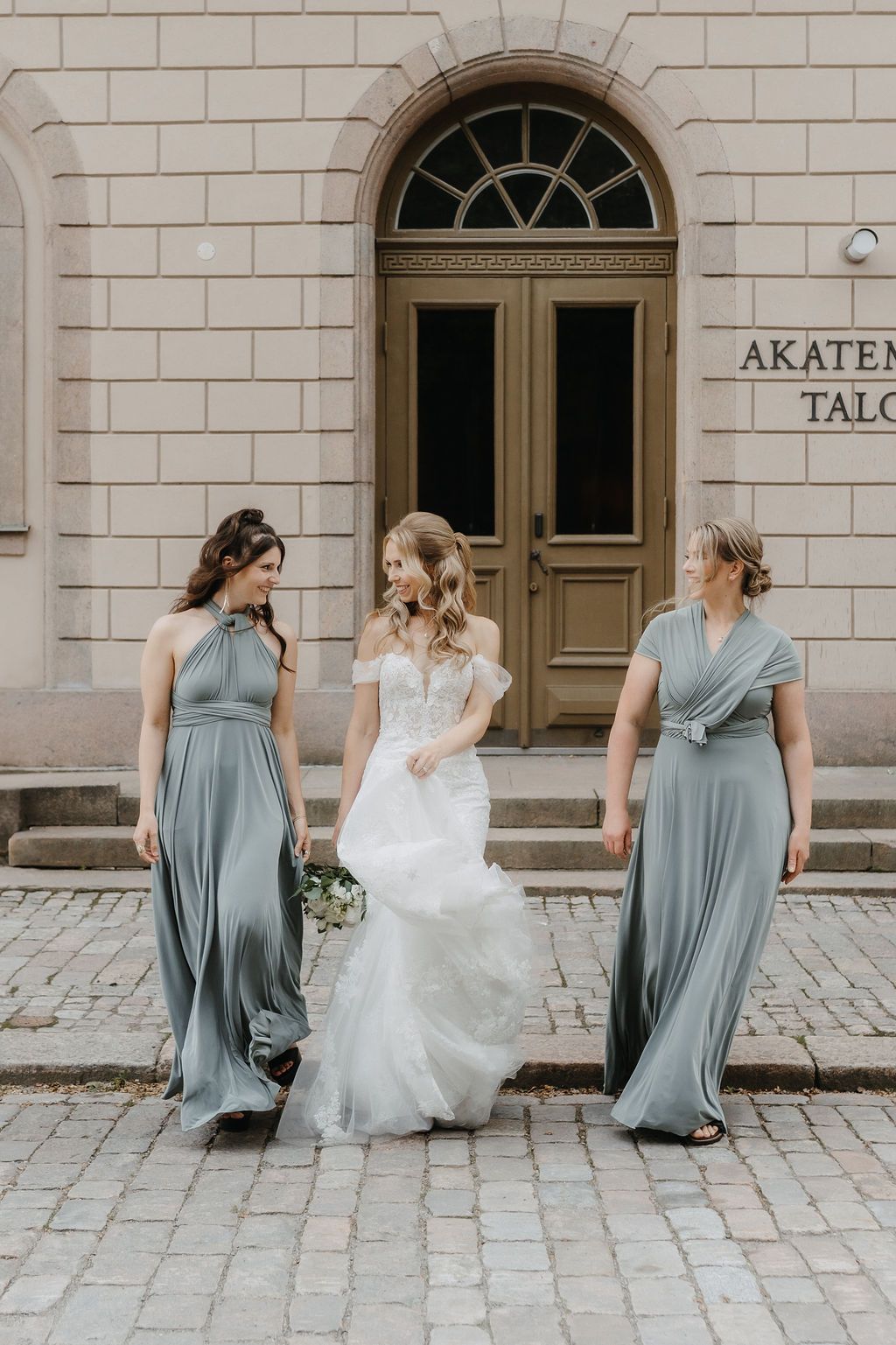 Bride and two bridesmaids in sage wrap dresses walk together outside Akatemia Talo on cobblestones.