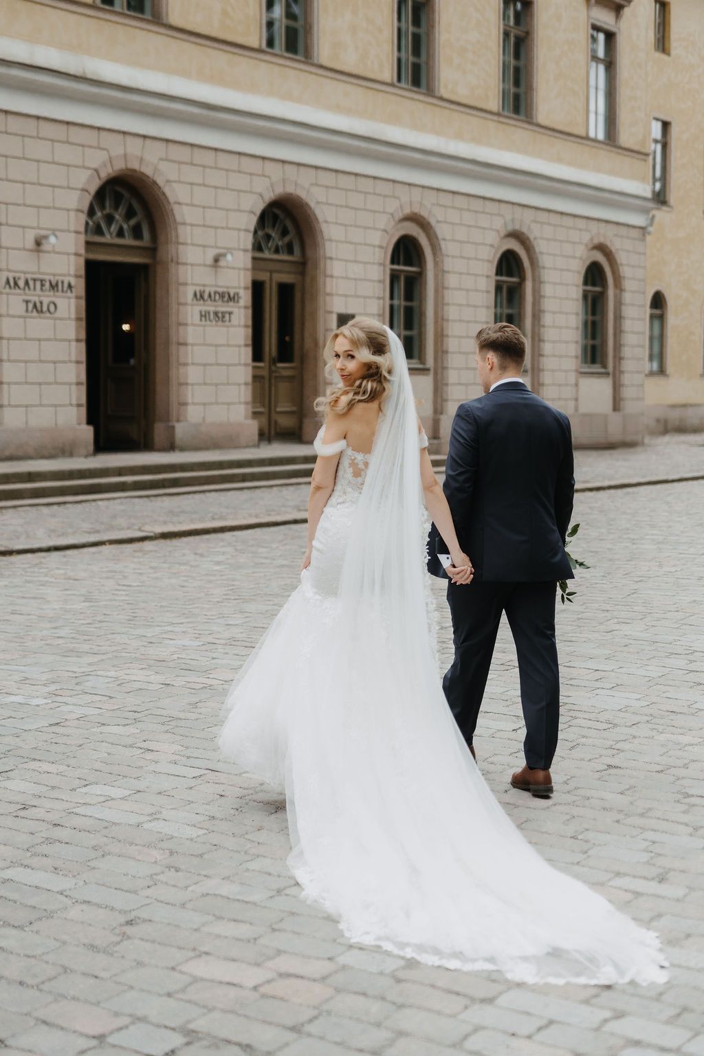 Bride in elegant lace gown with long veil holds groom's hand walking past Akademia Talo building.