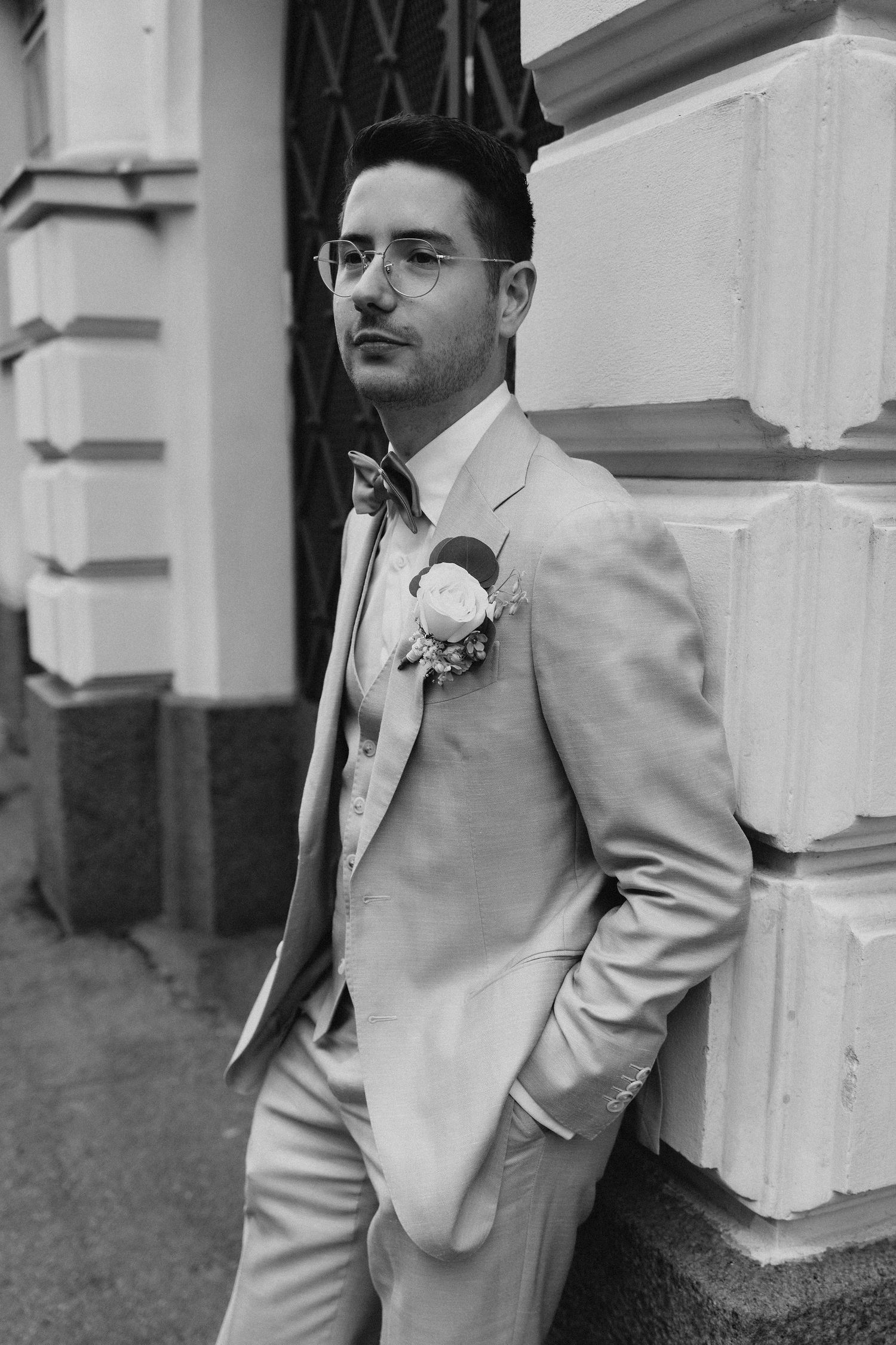 Groom in beige suit with boutonniere and glasses leaning against stone wall in black and white photo.