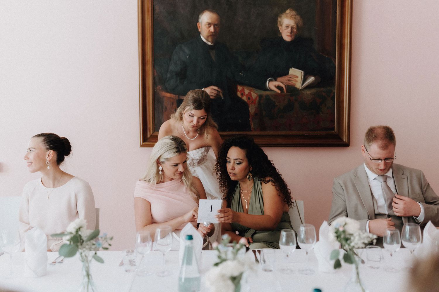 Wedding guests gather around a decorated reception table beneath a classic portrait painting indoors.