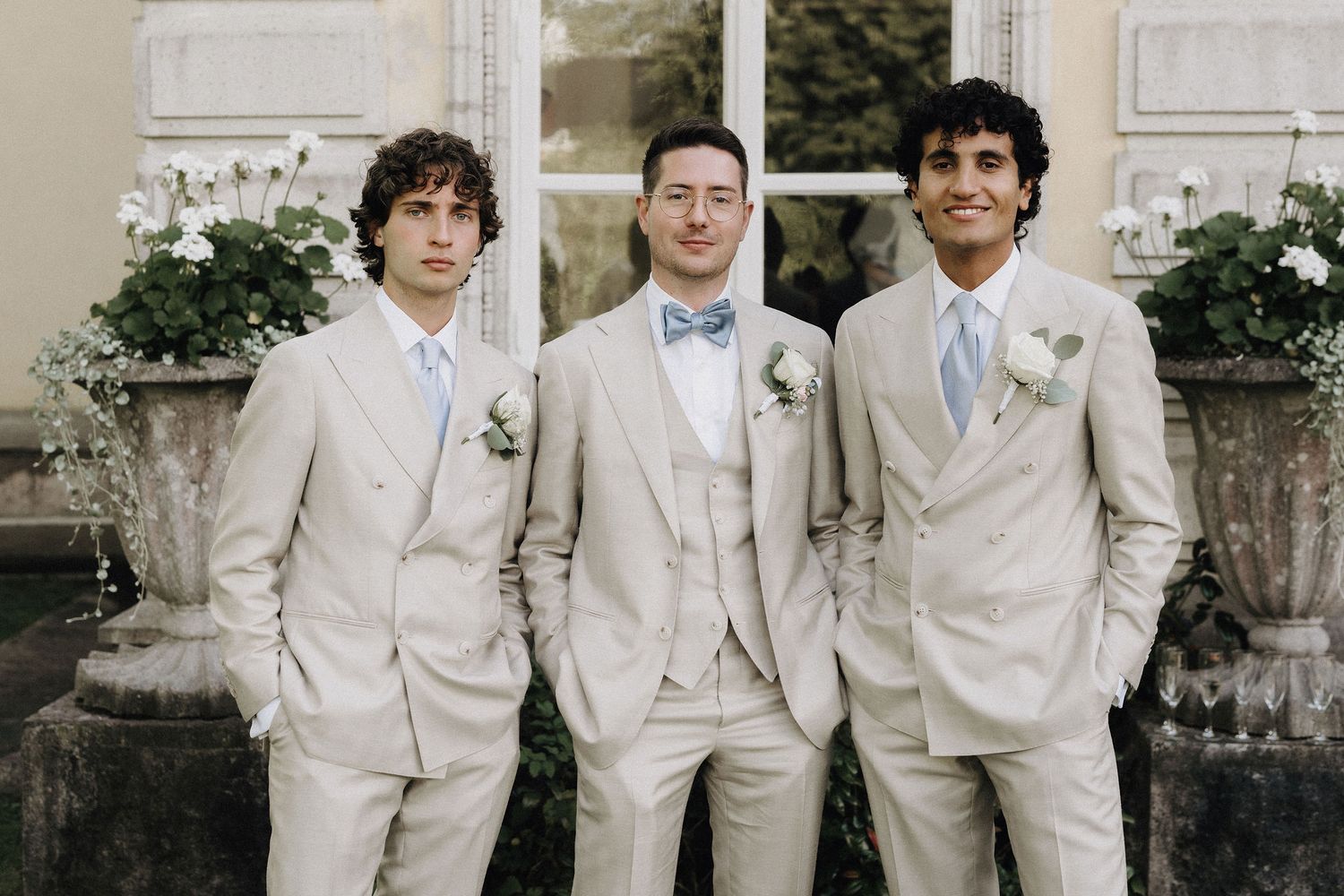 Three groomsmen in matching beige suits with boutonnieres posing outside a elegant building.