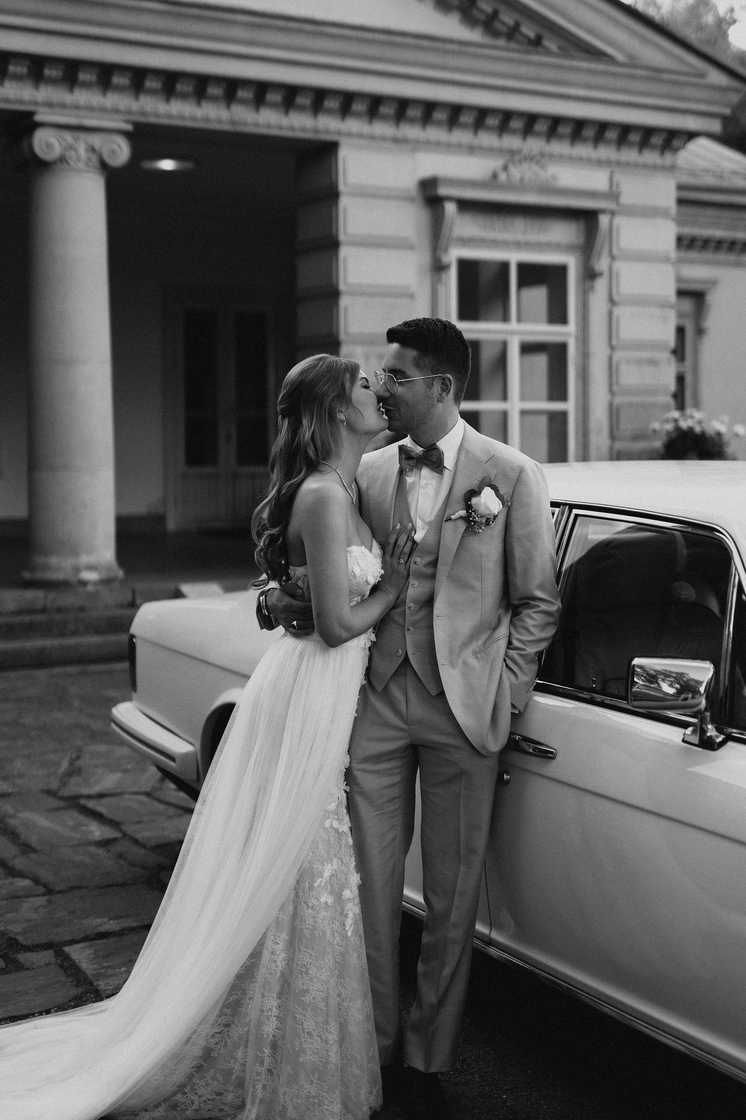 Black and white photo of bride and groom sharing a kiss beside a vintage car at a neoclassical venue.