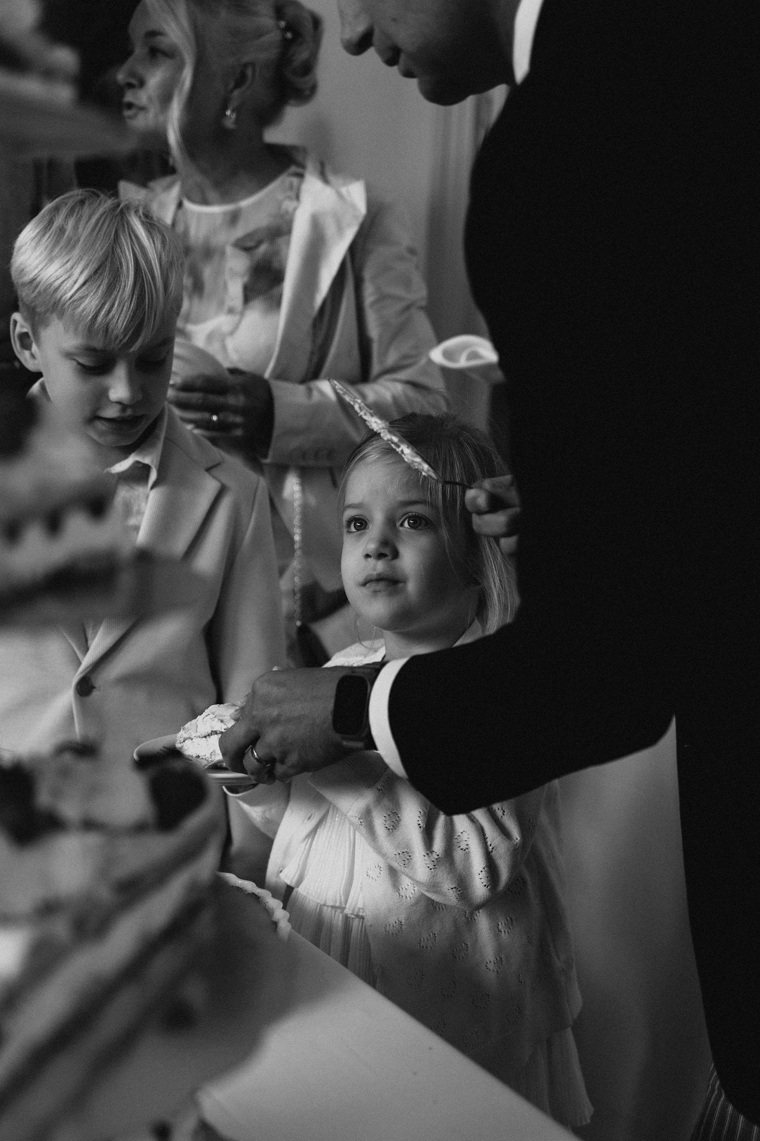 Black and white photo of a young flower girl receiving wedding cake at the reception table.
