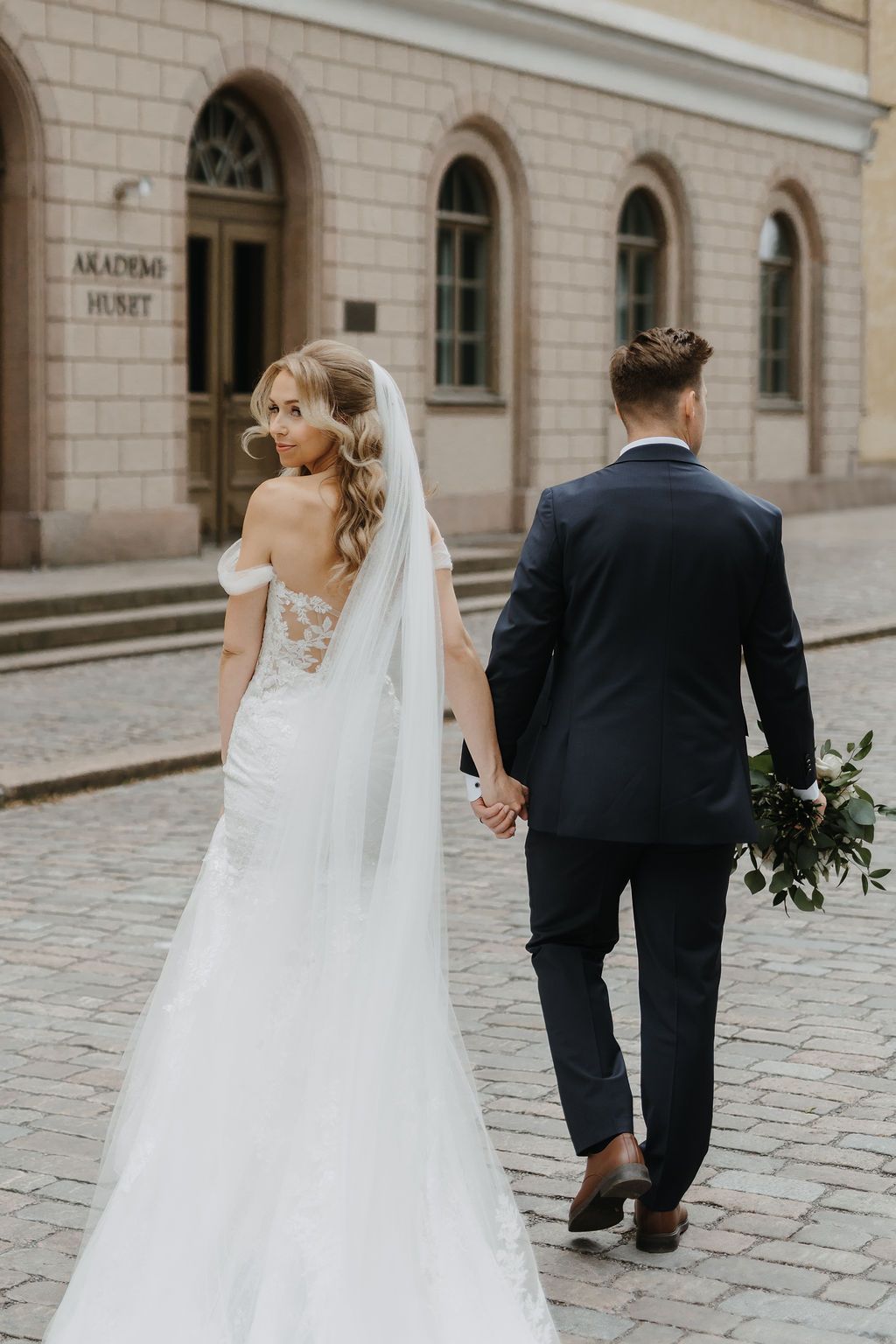 Bride in off-shoulder lace gown with cathedral veil walks hand in hand with groom on cobblestone street.
